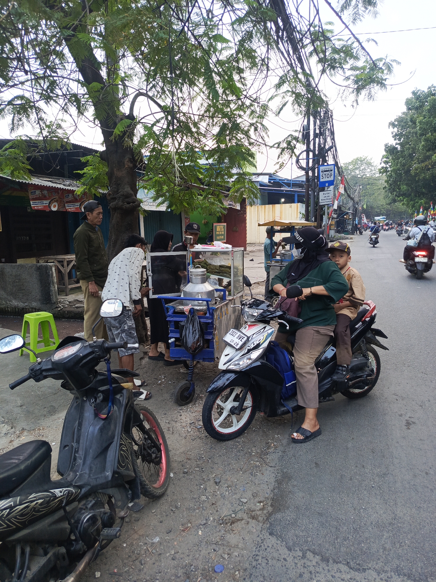 A street scene in West Jakarta, Indonesia, shows a group of people gathered around a food cart. A woman wearing a black hijab and a green shirt is seated on a black motorcycle, with a young boy in a brown shirt behind her. In front of the motorcycle is another black motorcycle parked on the side of the road. The food cart has a stainless steel container and a blue wheeled base, and two men are interacting near it. One man in a green jacket and tan pants stands to the left of the cart, while another man in a patterned shirt is leaning over it. Behind them, a building with a red and white awning is visible. The street is lined with trees, power lines, and other parked motorcycles and scooters, indicating a busy urban environment. A "STOP" sign is visible on a pole in the background.