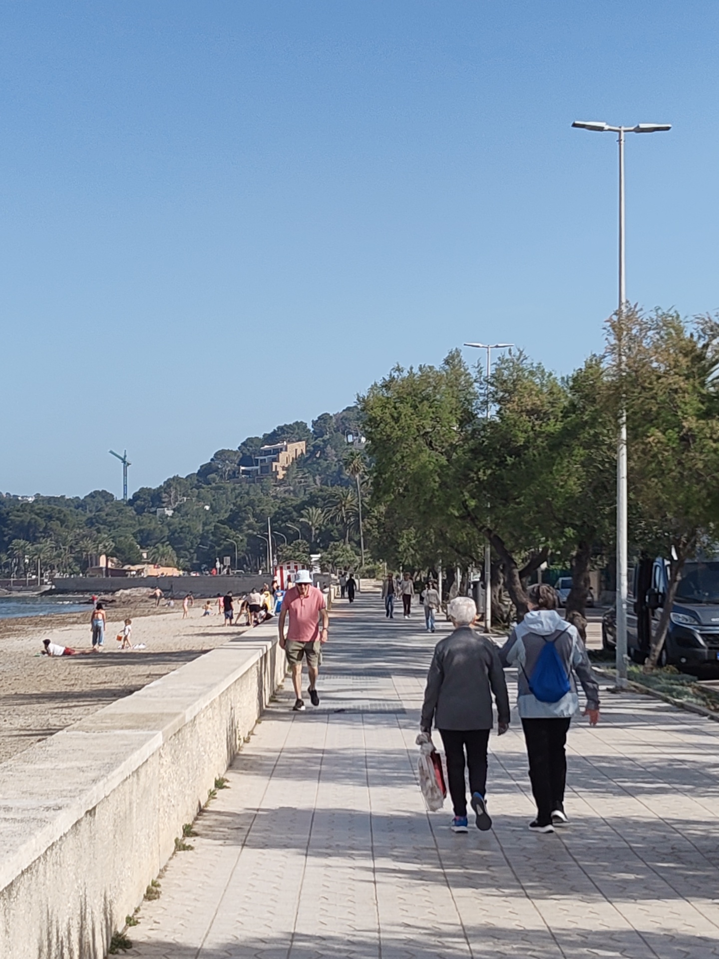 The image shows a promenade in Dénia, Spain, on a sunny day. Several individuals stroll along the paved walkway that runs parallel to the beach. On the beach, people engage in various activities, including sunbathing and playing. A low wall separates the promenade from the beach. In the background, a hill covered with trees is visible, with some buildings situated on its slopes. Streetlights line the promenade alongside trees, with vehicles parked alongside the far side of the walkway. The perspective is from behind two people walking away from the camera.