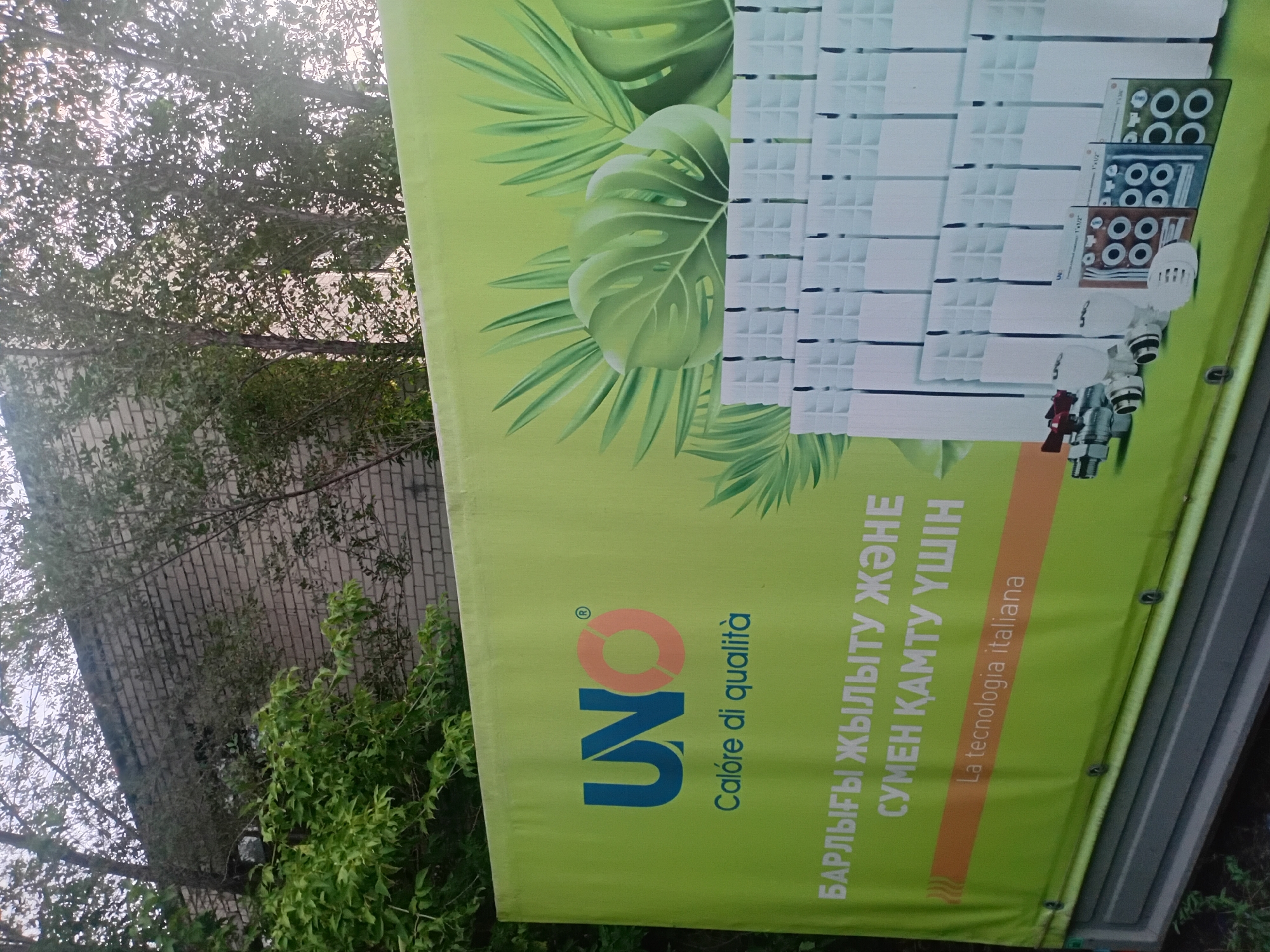 This image depicts a large, bright green billboard advertising heating radiators. The billboard is positioned outdoors, with trees and a brick building visible in the background. On the left side of the billboard is the logo "UNO" in blue and orange, with the Italian phrase "Calore di qualità" underneath, meaning "Quality Heat". To the right of the logo are stylized images of tropical leaves. The main visual on the billboard is a depiction of white radiators, with some plumbing fixtures and product packaging shown in the foreground. Text in what appears to be Kazakh is present on the lower left side of the billboard, reading "БАРЛЫҒЫ ЖЫЛЫТУ ЖӘНЕ СҮМЕН ҚАМТУ ҮШІН", which translates to "For all heating and coverage". Below this, in smaller font, is the Italian phrase "La tecnologia italiana", meaning "Italian technology". The presence of the "UNO" logo and the Italian text strongly suggests this billboard is advertising an Italian brand of heating products. Based on the text and the context of an advertisement for heating systems, it's plausible that this billboard is located in Semey, Kazakhstan, a city that experiences cold winters and would therefore have a market for such products. The billboard is mounted on a dark gray base. The image is taken from a slightly low angle, looking up at the billboard.