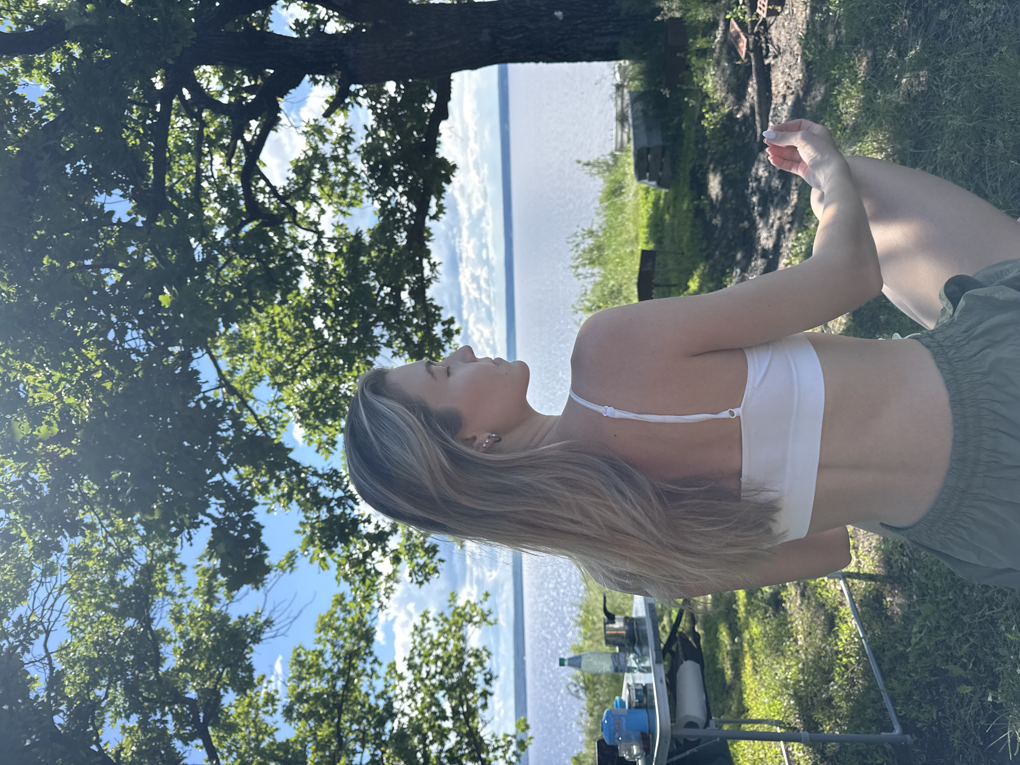 A medium, outdoor shot shows a woman seated by a body of water under a tree. The woman is wearing a white top and green shorts. She is facing left, with her head tilted back. Her right arm is raised. Behind the woman, there is a body of water, and some greenery. In the foreground, there is a small table with various items on it, including a blue propane tank, a bottle, and a roll of paper towels. The environment is a grassy area with a large tree to the left and a body of water in the distance. The location is the city of Unknown, Unknown.
