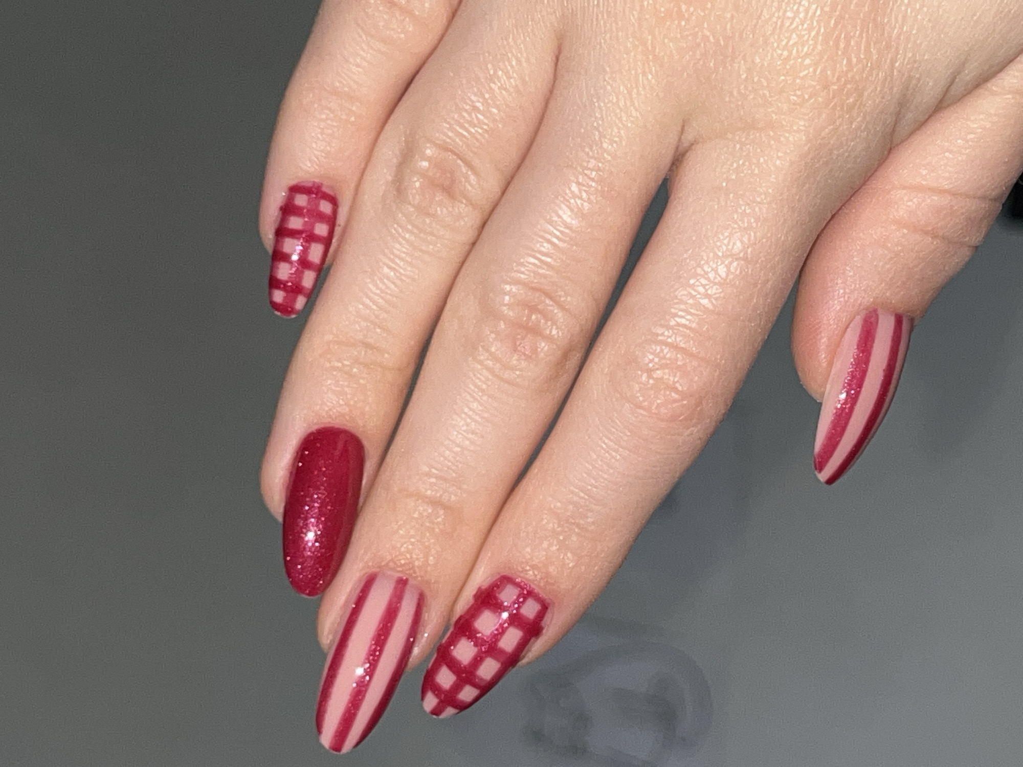 The media file features a close-up view of a person's left hand, showcasing a recently applied manicure. The hand is positioned against a plain, dark grey background, drawing focus to the elaborately designed fingernails. This scene takes place indoors, potentially in a nail salon or home in Perm, Russia.
Five long, shaped nails, possibly in a coffin or almond style, are visible. Each nail features a unique design within a cohesive color scheme of dark red/maroon and a light pink/nude shade. The nail on the thumb (partially visible on the right) and the ring finger display vertical stripes, where dark red/maroon lines are painted over a pink/nude base, accented with a subtle glittery finish.
The index and pinky fingernails are adorned with a plaid or grid pattern, rendered in the dark red/maroon color over a pink/nude base. In contrast, the middle fingernail is a solid dark red/maroon, distinguished by its prominent glittery finish.
No specific activity is depicted beyond the presentation of the manicured hand. The indoor setting offers no further location details, time of day cues, or visible text within the image.