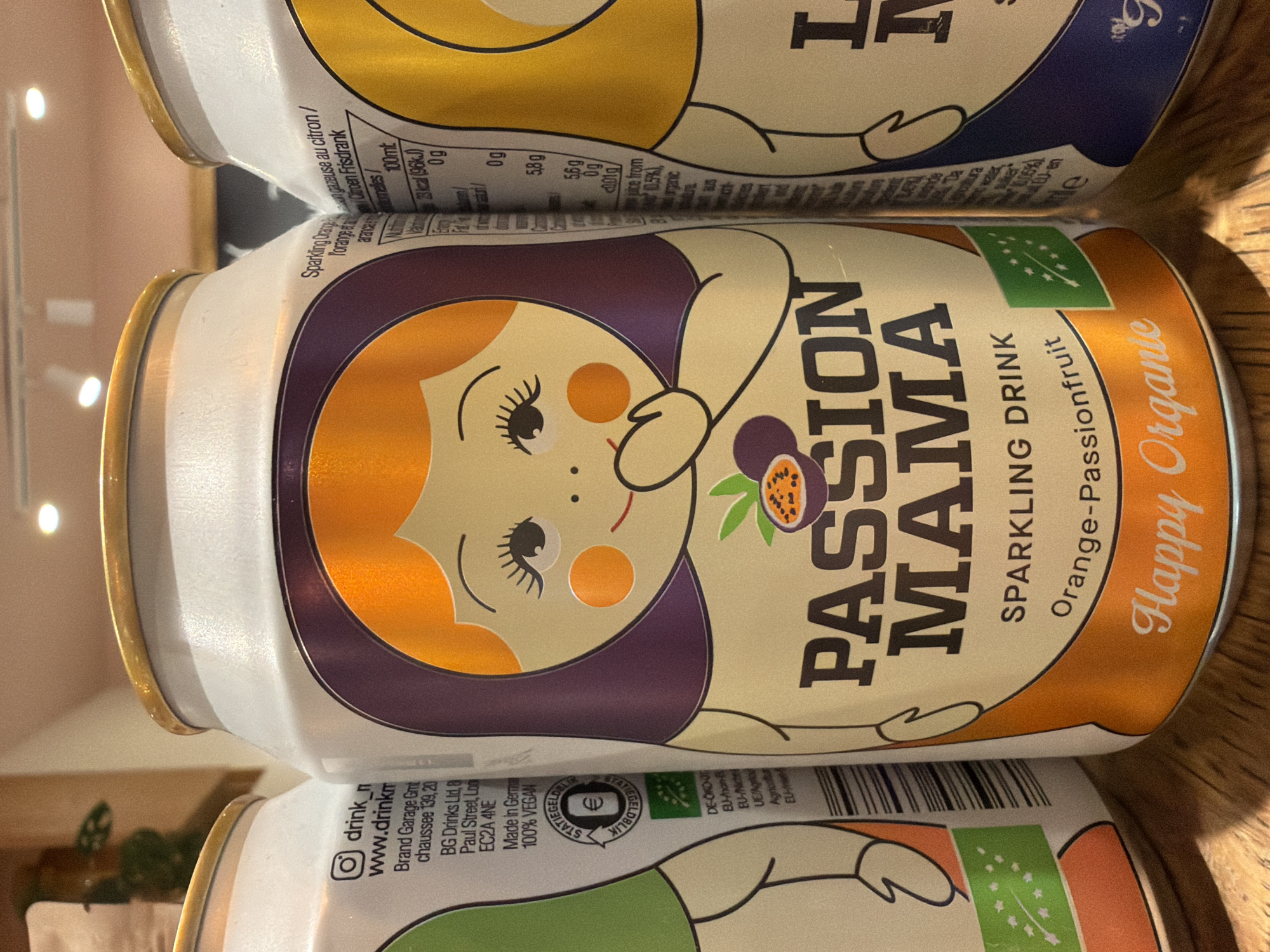 A close-up shot depicts a can of "Passion Mama" sparkling drink, specifically the "Happy Organie" Orange-Passionfruit flavor. The can features a cartoonish female figure with orange hair, a purple dress, and orange cheeks. The words "Passion Mama" are prominently displayed in bold, capitalized letters next to an illustration of a passion fruit. The can is labeled "Sparkling Drink" and "Orange-Passionfruit" with "Happy Organie" in a stylized, flowing script. A green EU organic certification symbol is visible. Other cans of similar drinks are partially visible above and below the main can, suggesting they are stacked. Some text on the cans below the main can are partially visible, detailing ingredients and nutritional information.
