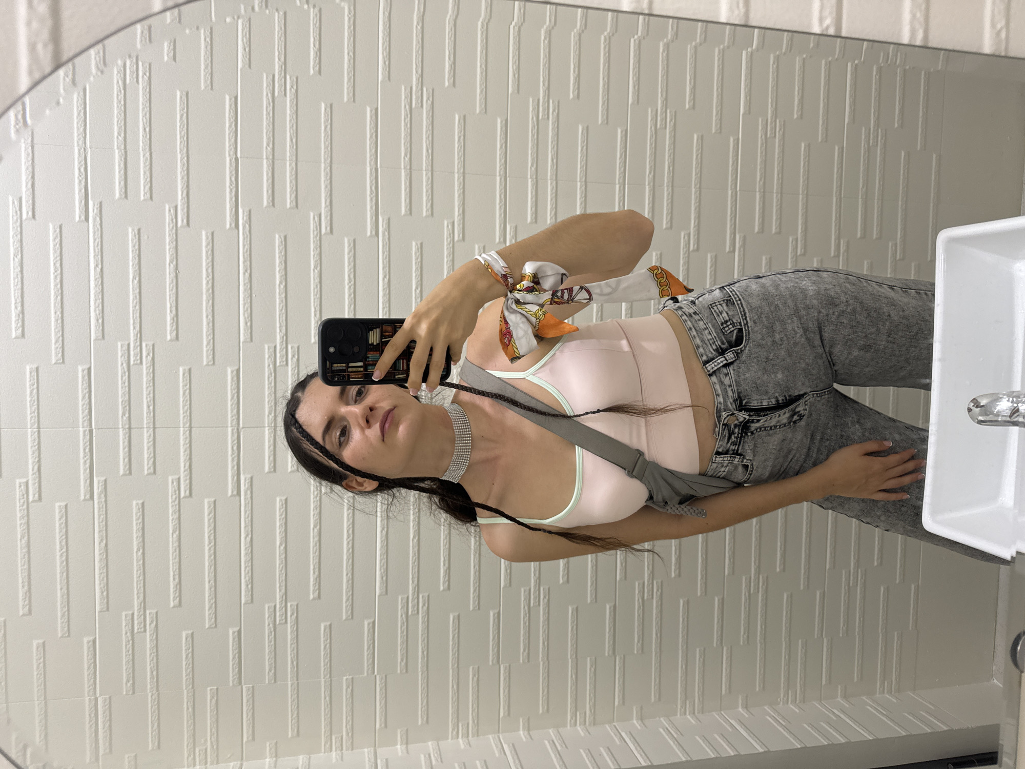 A young woman is taking a selfie in a bathroom. She is wearing a light pink top with a light green trim, grey acid wash jeans, and a sparkling choker. A colorful scarf is tied around her arm. She has dark hair, with two small braids framing her face. She is holding a smartphone with a bookcase design case. The bathroom has white textured rectangular wall tiles. Part of a white sink is visible on the right. The location is inferred to be in the Phu Thuy Ward, Vietnam, based on the query.