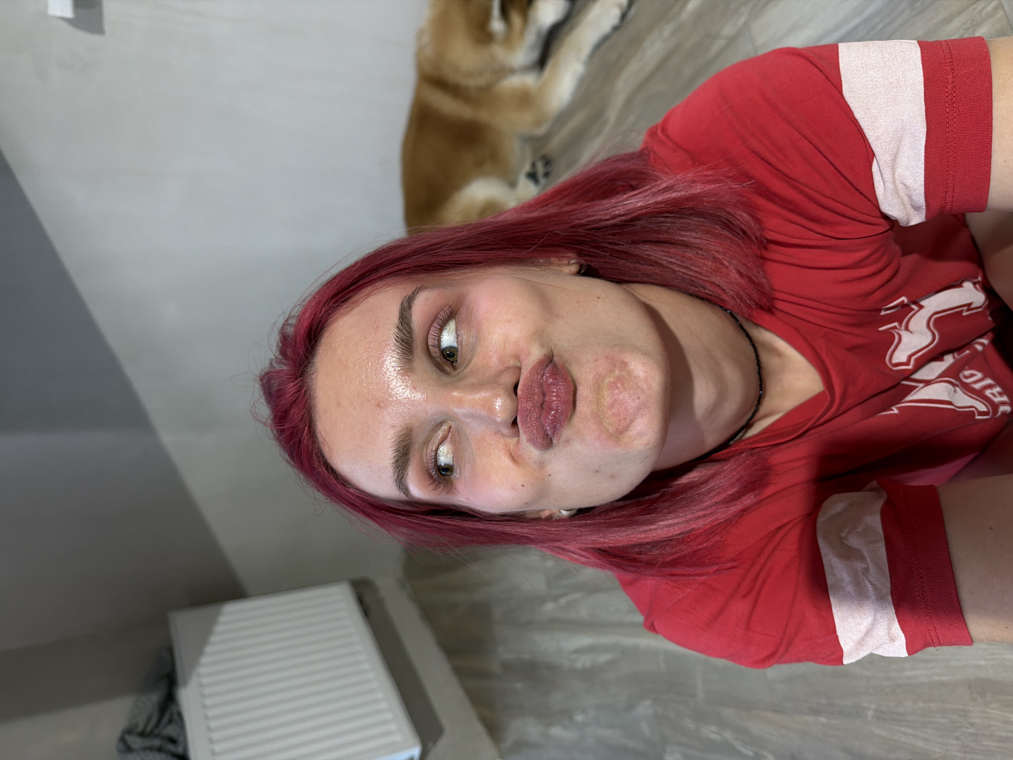 A young woman with bright pink hair is making a kissing face at the camera, her eyes looking to the side. She is wearing a red t-shirt with white stripes on the sleeves and a black choker necklace. Her makeup includes eyeshadow and mascara. Behind her, a dog is lying down on a wooden floor. The background appears to be an indoor space with a radiator and a grey wall. The image's context, specifically its relation to Chaikovsky, Russia, is not discernible from the visual information presented.