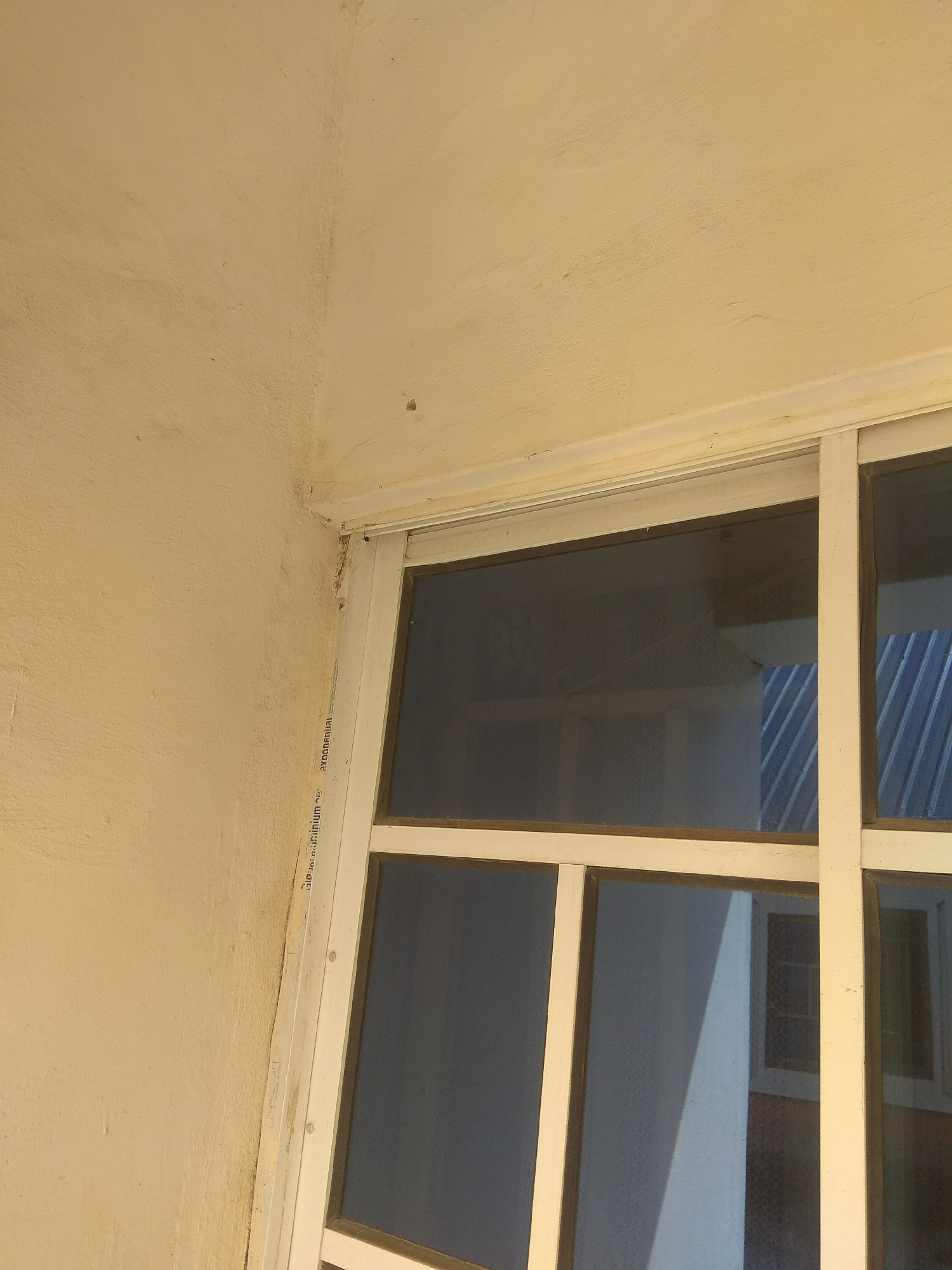 The image shows a close-up of an exterior wall with a window frame. The wall is painted a light yellow and appears to have some texture and minor imperfections. The window frame is white and has multiple panes. The glass in the panes is dark and reflects the surroundings, including a corrugated metal roof structure. Some writing is visible on the vertical part of the window frame, but it is partially obscured and difficult to read clearly. The lighting suggests it might be daytime, possibly overcast or in shadow, given the lack of strong direct sunlight. The overall scene depicts a part of a building's exterior, likely in a residential or commercial setting. No people are visible in the frame.