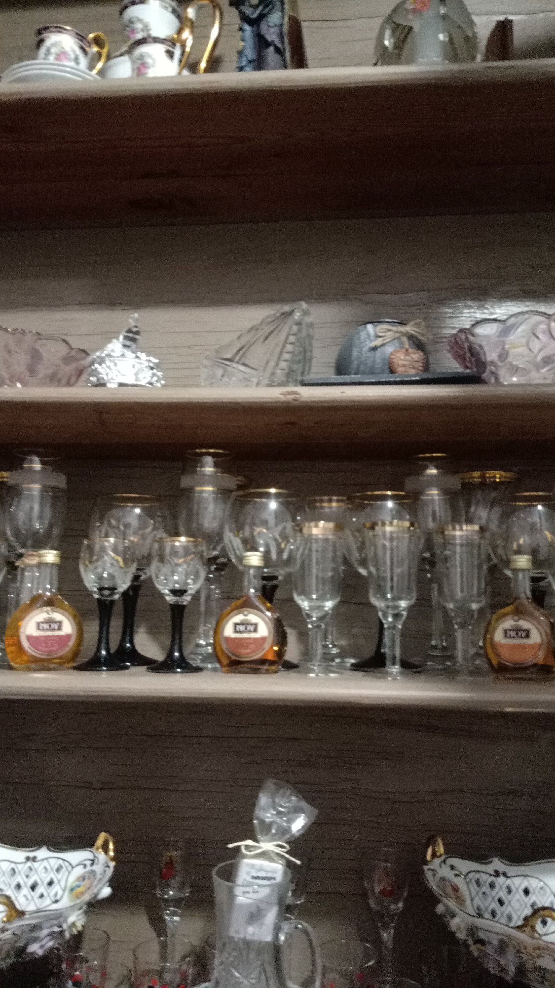 The image shows a wooden shelf containing various glassware and decorative objects. On the top shelf, there are teacups and saucers, along with other decorative items. The second shelf holds glass bowls and decorative pieces. The third shelf displays an assortment of wine glasses and miniature liquor bottles. The bottom shelf contains decorative glass bowls and more glassware. The environment appears to be a home setting. There's no indication of a specific location within Yerevan, Armenia, but the "NOV" liquor bottles suggest a possibility of Armenian origin.