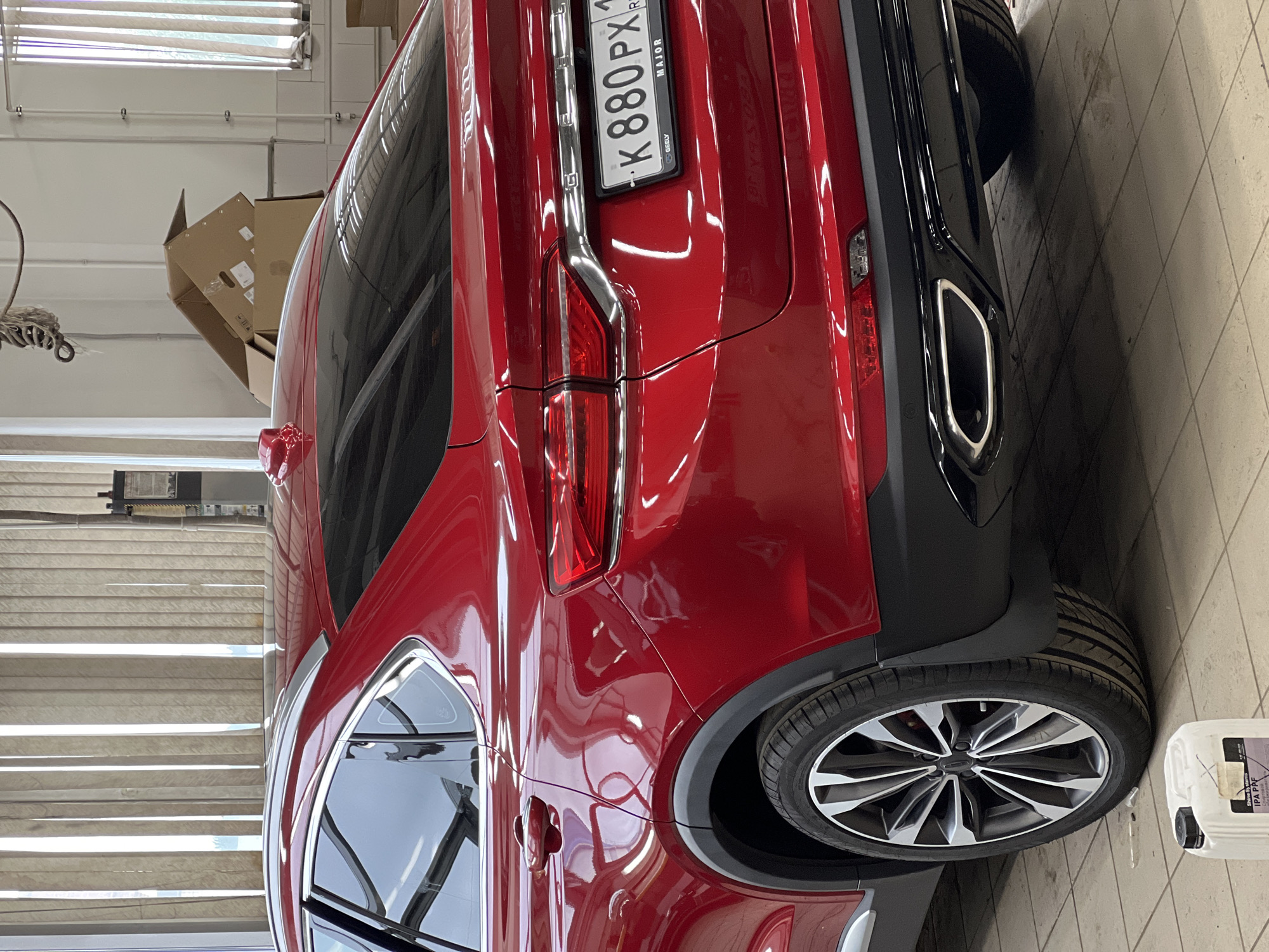 A red Geely Tugella (also known as Xingyue) coupe SUV is positioned indoors within what appears to be a car detailing or service center. The vehicle exhibits a highly reflective finish on its metallic red paintwork. It is equipped with a Russian license plate, "K 880 PX 1", which includes the "RUS" country identifier. A dealership frame beneath the plate displays "MAJOR" and "GEELY". The region code "1" on the license plate corresponds to the Republic of Adygea, Russia. Visual information within the image does not provide identifiable cues to confirm the requested location of Izhevsk, Russia.
The facility's floor is covered with light-toned square tiles. Natural light illuminates the space, entering through tall windows fitted with vertical blinds. In the background, several cardboard boxes are stacked, and a vertical metal structure, possibly a component of a vehicle lift or workshop apparatus, is present, with coiled wires or ropes suspended above it. On the floor near the vehicle's rear-left wheel, a white plastic container with a black cap is visible, partially labeled "Shine Systems" and "IPA PPF", indicating the use of isopropyl alcohol for paint protection film applications. The car features multi-spoke alloy wheels and a black rear bumper fascia incorporating a chrome-edged exhaust outlet on the right side. No individuals are depicted in the scene.