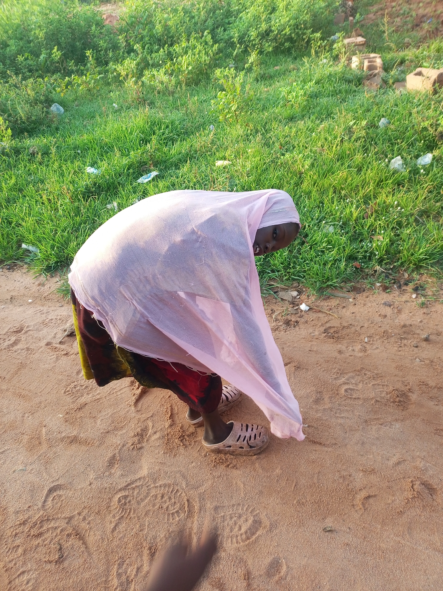 A young Black female is bent over on a sandy path with grass and debris in the background. She is wearing a pink hijab and a colorful skirt, and appears to be wearing sandals. The lighting suggests it is daytime. The environment is outdoors with natural vegetation and a sandy surface. There is debris in the grass.