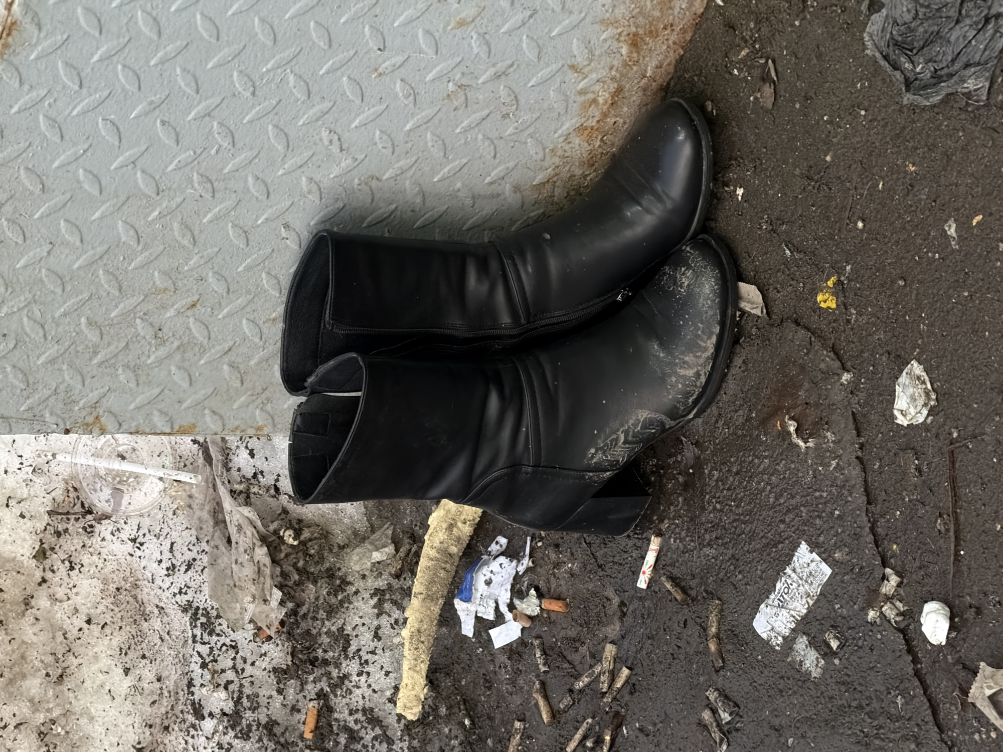 The scene depicts a pair of black, mid-calf women's boots with chunky heels and interior zippers, positioned upright against a light gray diamond-plate metal surface. The immediate ground consists of dark, wet soil and patches of dirty, melting snow. The left boot's toe and side are significantly soiled with mud. The environment is heavily littered with numerous discarded cigarette butts, fragmented white and blue paper, a clear plastic cup containing a white straw partially submerged in the dirty snow, and a segment of yellowish foam insulation. The metal surface exhibits rust staining at its lower right section. The location is an urban outdoor environment in Khimki, Russia.