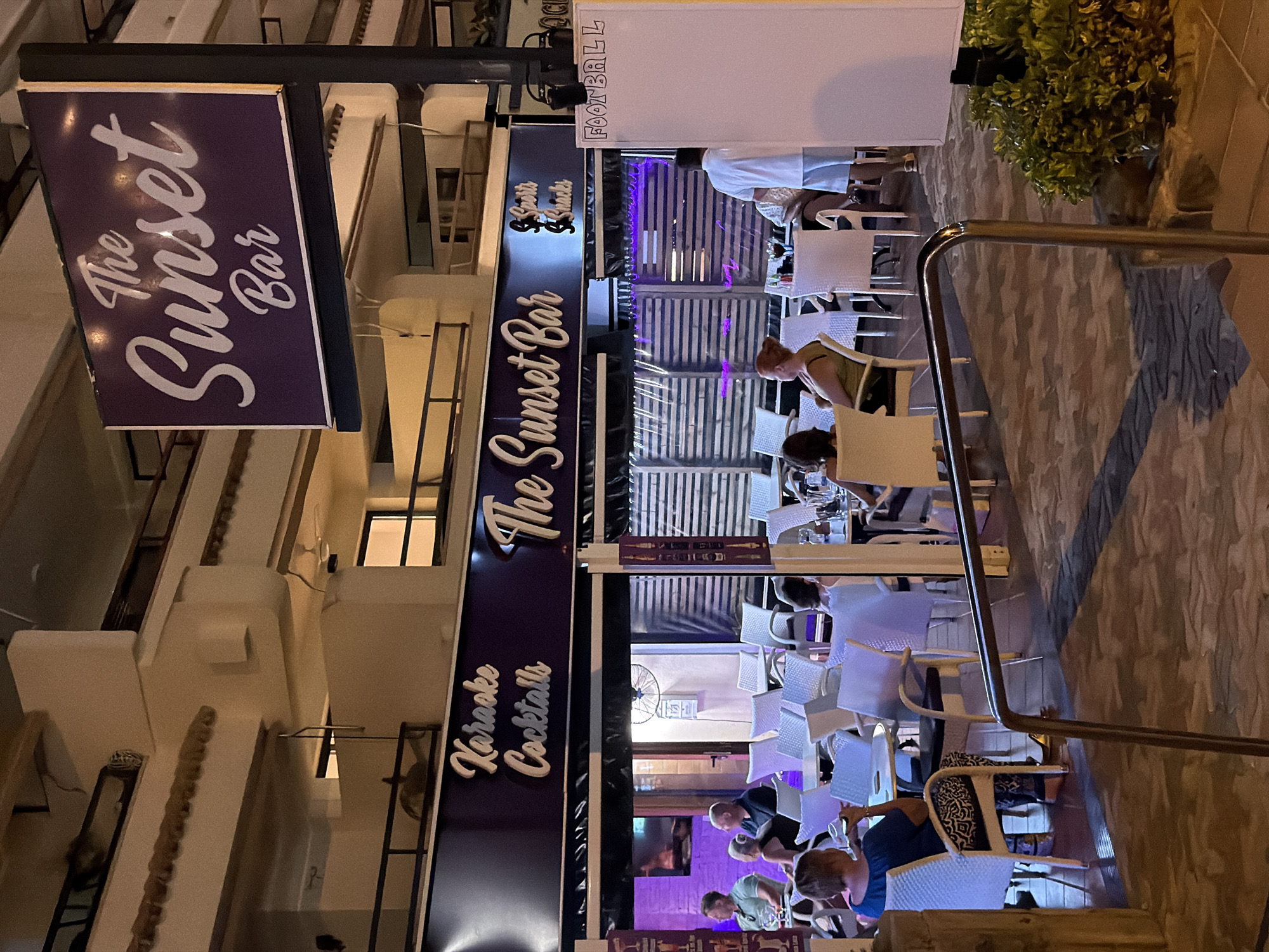The image depicts the exterior of a bar named "The Sunset Bar" located in Palmanova, Spain. The bar has a prominent sign with the name in stylized white lettering against a dark purple background. Below the main sign, another smaller sign indicates "Karaoke Cocktails". Further to the right, there's a sign that reads "Sports & Snacks".
The bar features an outdoor seating area with numerous white tables and chairs. Some patrons are visible seated at these tables, engaged in conversation or appearing to dine and drink. The area is covered by a dark awning, and protective clear curtains are drawn on the sides, suggesting it is evening or night. Inside, some areas are illuminated with purple lighting.
To the right of the establishment, a white board with "FOOTBALL" written in black marker is visible, indicating that sporting events might be shown. A man in a white t-shirt and blue shorts stands near this board. The overall atmosphere suggests a relaxed evening setting at a bar or restaurant. The pavement outside has a decorative tiled pattern.