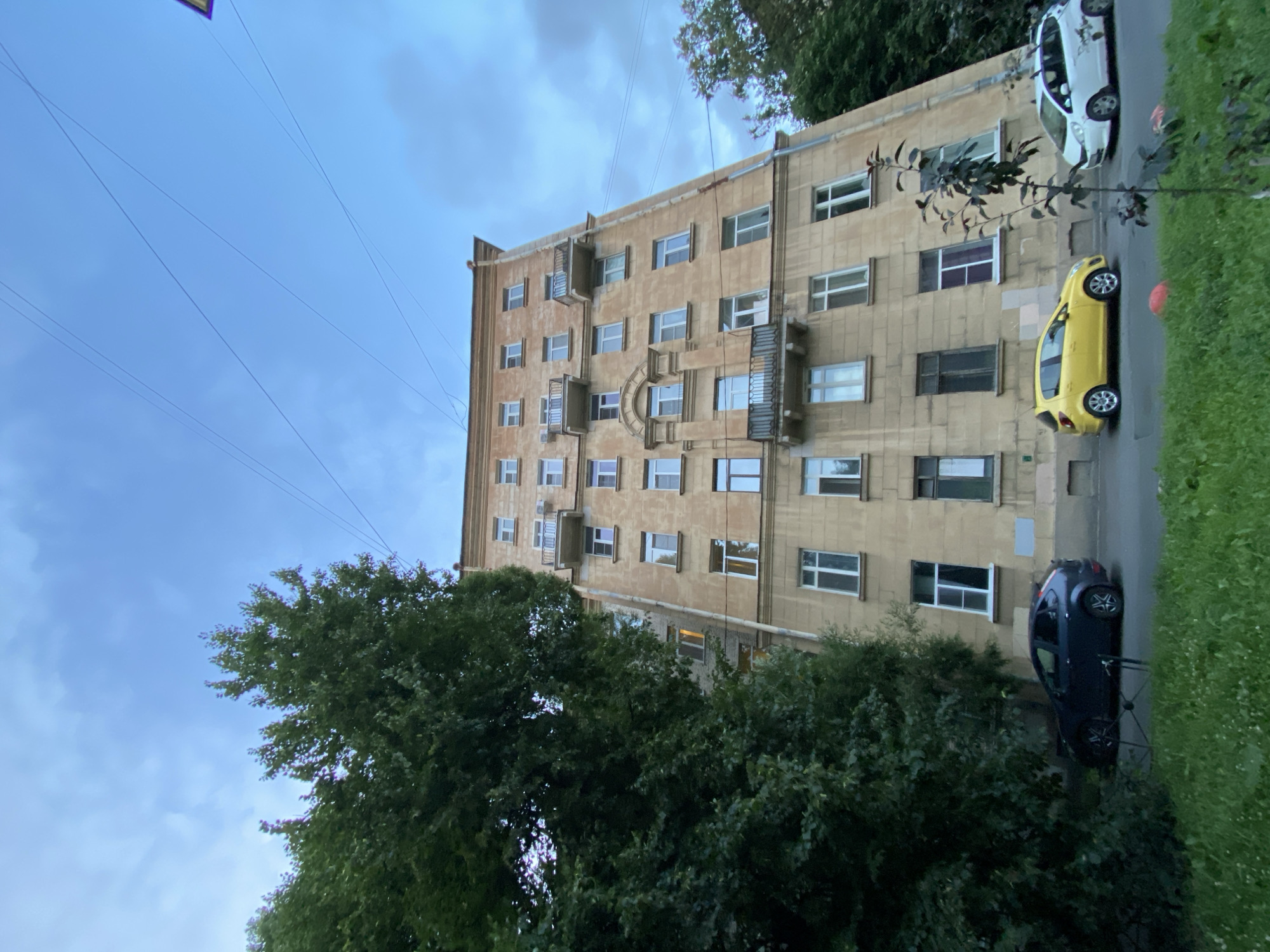 The image depicts a street view in Saint Petersburg, Russia, showcasing a tall, multi-story building with a beige facade, several windows, and balconies. The building appears to be an apartment complex. In front of the building, there are three parked cars: a dark blue hatchback on the left, a bright yellow hatchback in the middle, and a white station wagon on the right. The foreground shows a grassy area with some trees, indicating a residential or park-like setting. The sky above is partly cloudy and blue. The perspective of the shot is low-angle, looking up at the building and trees.