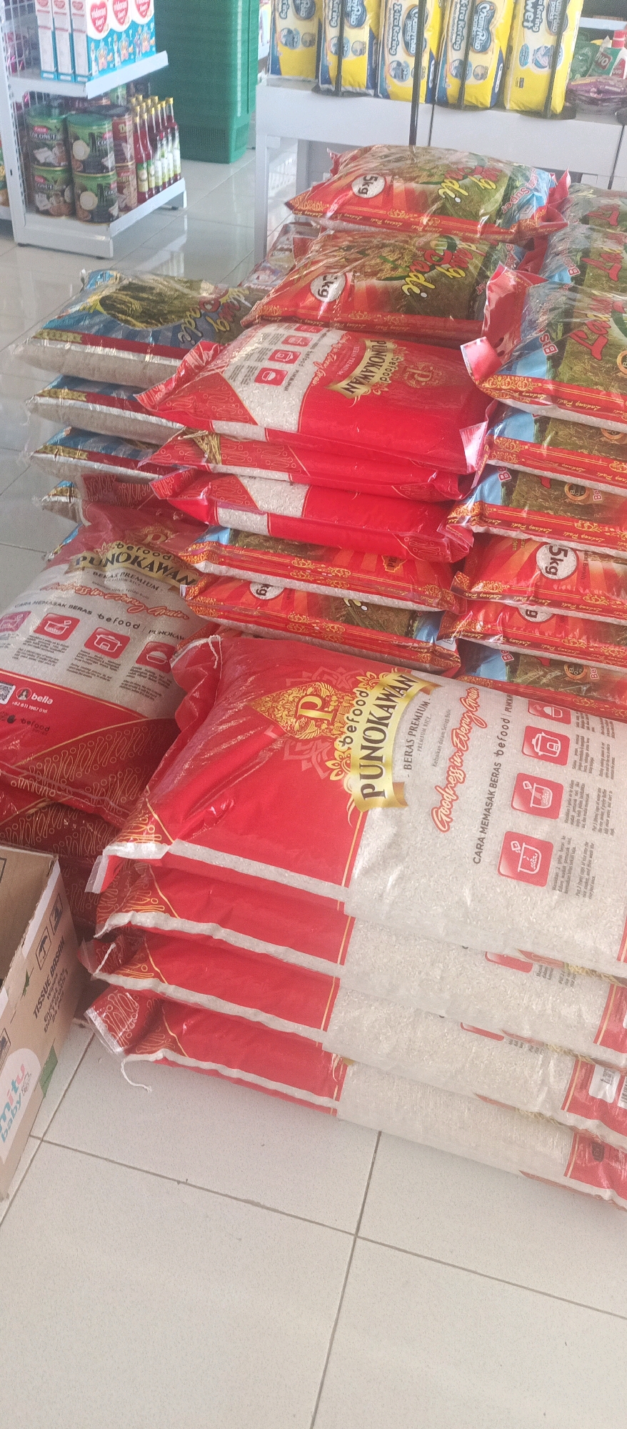 The image depicts a retail environment, likely a supermarket or convenience store, in Lamongan, Indonesia. The foreground is dominated by stacks of bagged rice, predominantly branded "befood PUNOKAWAN" and "Rasa Premium". These bags are red and gold and some are labeled with "5kg". To the left, a cardboard box with "MITU BABY" branding is partially visible. In the background, shelves are stocked with various products, including what appear to be canned goods, bottled beverages, and packaged snacks. The flooring is tiled. The overall impression is one of a well-stocked retail display, with a focus on staple food items.