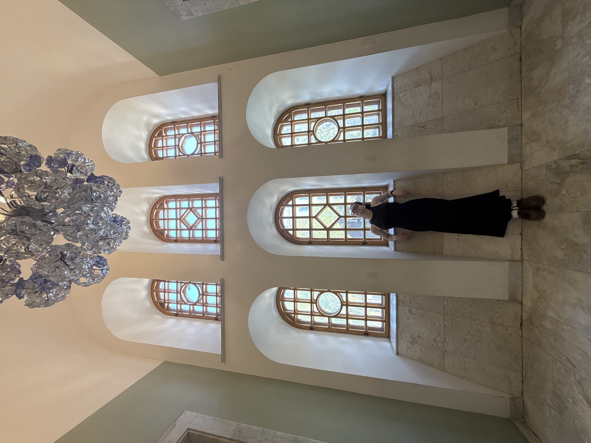 The image shows the interior of an unidentified building in Moscow, Russia, featuring three rows of arched windows with lattice-like frames. A woman in a long black dress and brown boots is leaning against the wall between the second and third windows on the right side. A large chandelier is positioned in the upper left of the frame. The walls are primarily off-white with a light green section near the top. The floor and wall adjacent to the windows are tiled with a light-colored stone.