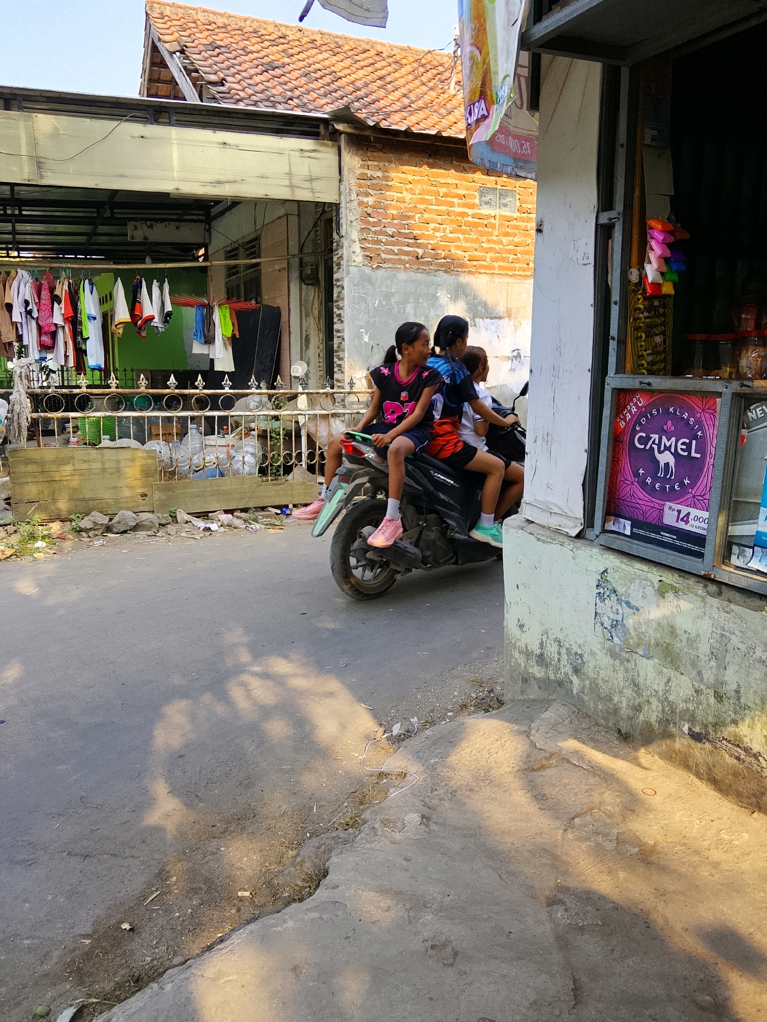 The image shows a street scene in Cirebon, Indonesia, likely in a residential or mixed-use area. In the foreground, a motorcycle is moving on a paved road. Three young people are riding the motorcycle: two girls are seated at the back, and a third person, whose gender is not clearly discernible, is in the middle. The girl closest to the viewer has dark hair pulled back in a ponytail and is wearing a dark shirt with a pink design and dark shorts. The girl behind her has dark hair and is wearing a blue and white t-shirt. They are all dressed casually.
The motorcycle is a dark color, and the rear wheel and part of the fender are visible.
In the background, there is a building with a red tile roof and brick walls. A section of the building appears to be a small storefront or kiosk, with items displayed behind a window, including a poster advertising "Camel Classic Kretek" cigarettes, priced at RP 14.000. Clothes are hanging on a clothesline in front of another part of the building, suggesting a domestic setting. A metal fence with decorative elements runs along the side of the property. The environment appears somewhat cluttered, with debris and rocks on the side of the road and in front of the fence. Sunlight casts shadows across the road and the foreground.