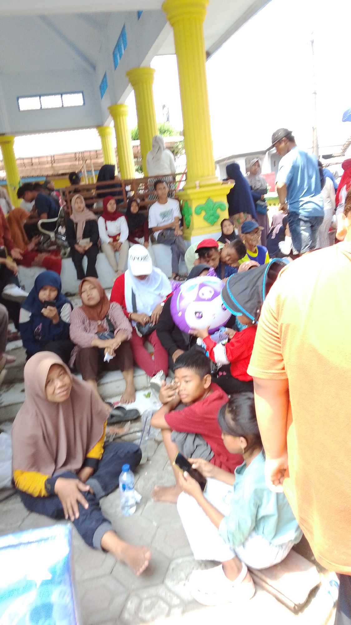 The image depicts a gathering of people, possibly at an outdoor event or gathering space, in Mancar, Indonesia. The scene is characterized by a bright, sunny atmosphere with a structure featuring yellow pillars and a white ceiling overhead, suggesting a pavilion or covered seating area.

In the foreground, several individuals are seated on steps and the ground. A woman wearing a light brown headscarf and a yellow and brown top is on the left, with her legs crossed. Next to her, a person in a dark blue jacket and headscarf is looking down at their phone. To their right, a woman in a maroon headscarf and brown top is seated, holding her hands in her lap. A child in a red shirt is seated in front of them, leaning forward and appearing to engage with something.

Further back, a group of people are seated on elevated steps. A woman in a white headscarf and red top is prominent in the center, and a person in a blue shirt and cap is standing to the right, looking away from the camera. A purple balloon with a cartoon face is held by someone in the crowd. Other individuals, dressed in various casual attire, are visible in the background, some standing and some sitting, suggesting a community event.

The ground is paved with hexagonal tiles, and a plastic water bottle is visible near the woman in the brown headscarf. The overall impression is of a casual social occasion, with people of different ages interacting in a public space in Mancar.