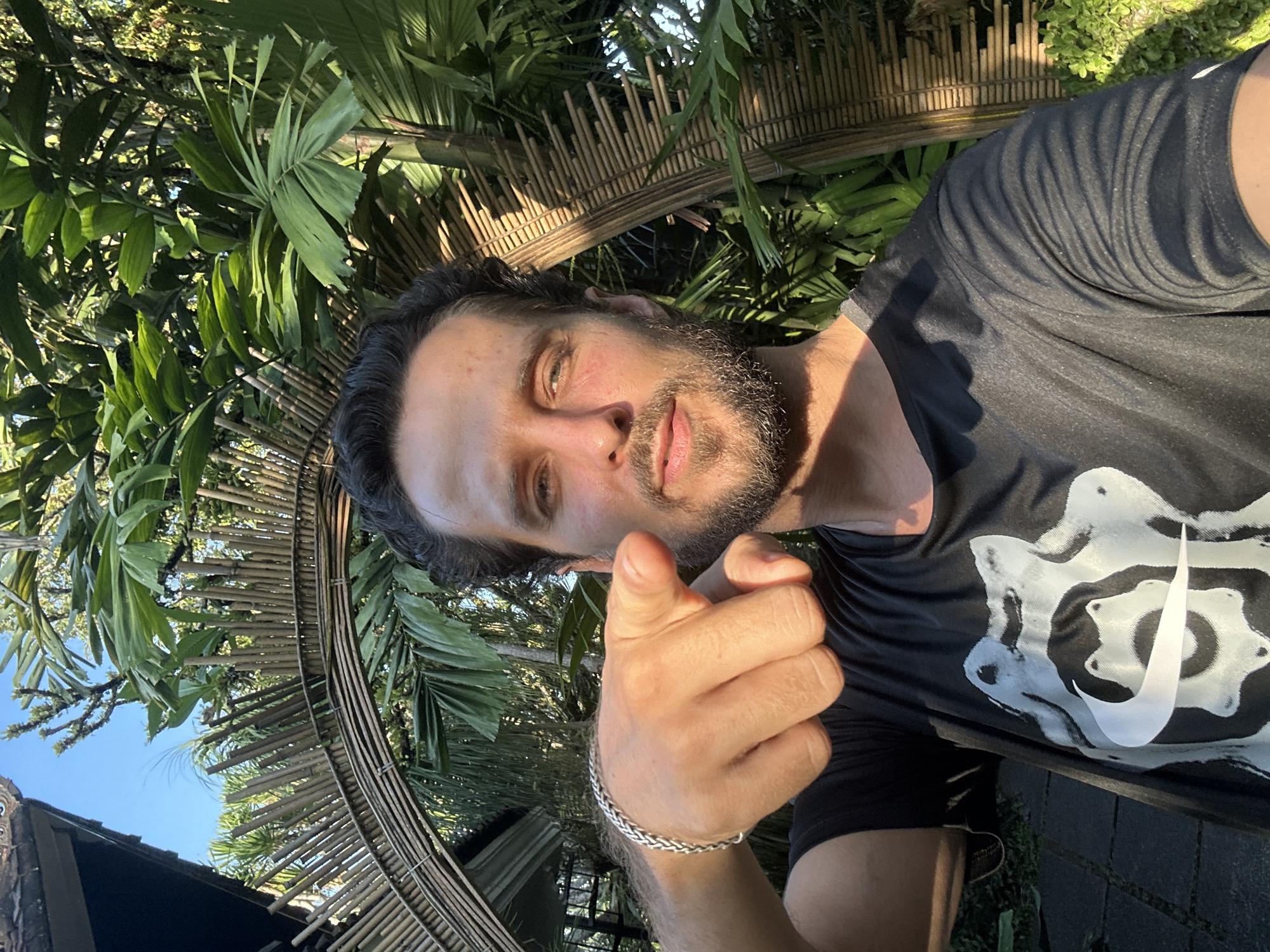 A man with dark hair and a beard points directly at the camera while holding up his right hand. He is wearing a black t-shirt with a white Nike logo and a silver bracelet on his left wrist. The background is filled with lush green palm fronds, a bamboo fence, and a clear blue sky, suggesting a tropical outdoor setting. The lighting indicates it is daytime, possibly late afternoon due to the warm, golden hue on the man's face and the shadows. The overall mood is energetic and engaging, as if the man is interacting with the viewer. The scene appears to be in a tropical outdoor area, with the presence of dense foliage and natural bamboo structures.