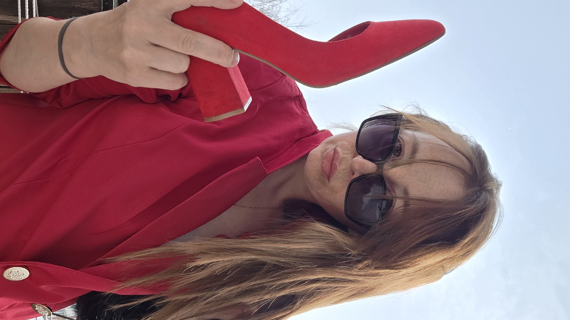 A female subject is depicted in a chest-up, outdoor photograph under a bright, clear sky. Her long, reddish-brown hair is visible, along with freckles on her nose and cheeks. She wears dark, rectangular sunglasses and has pursed lips. The subject is dressed in a red blazer with lapels and gold-colored, embossed buttons on the right side. A thin gold necklace is visible around her neck, and a dark band is present on her left wrist. She holds a single red high-heeled shoe with a pointed toe and a red block heel in her left hand. The background shows faint, bare tree branches on the left and a partial dark railing at the lower left. The image does not contain specific visual identifiers for a precise location within the city of Chaikovsky, Russia.