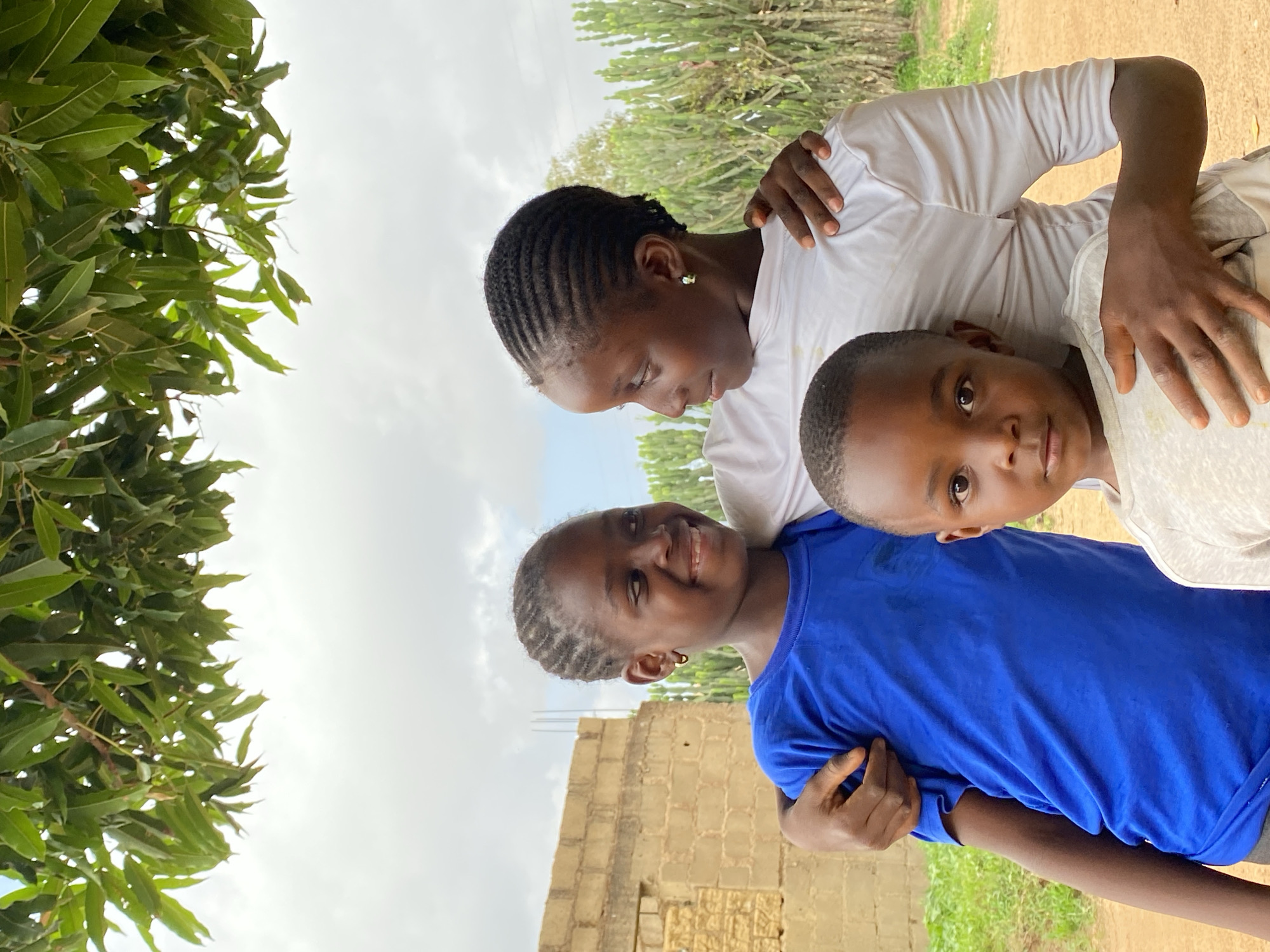 The image shows three individuals in an outdoor setting, likely in Kuru Dungus, Nigeria. The three people, a woman, a girl, and a boy are closely huddled together.
The woman is positioned at the top of the image. She has dark skin, short black braided hair, and is wearing a white shirt. She is looking down towards the other two individuals. The girl, wearing a blue shirt, is smiling and is situated at the bottom of the image. Next to her is a boy in a white shirt, looking at the camera.
The background includes a building constructed from tan-colored blocks and foliage. The sky is partially visible with cloudy conditions. The setting appears to be a rural area. The image seems to be rotated.