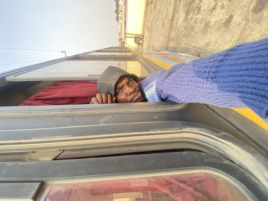 An adult male is observed leaning out of an open window of a stationary passenger bus, positioned on a paved surface in Issele Uku, Nigeria. The individual, of dark complexion, wears a dark bucket hat and a blue knit sweater. He grips the dark-colored window frame with his right hand, his head and shoulders visible from the chest up. His gaze is directed towards the camera with a serious expression. Red curtains are visible inside the bus through the window opening. The bus exterior features yellow and light blue sections. The surrounding environment consists of a bright, clear sky, and in the background, several light-colored multi-story buildings with dark roofs are visible, along with overhead power lines.