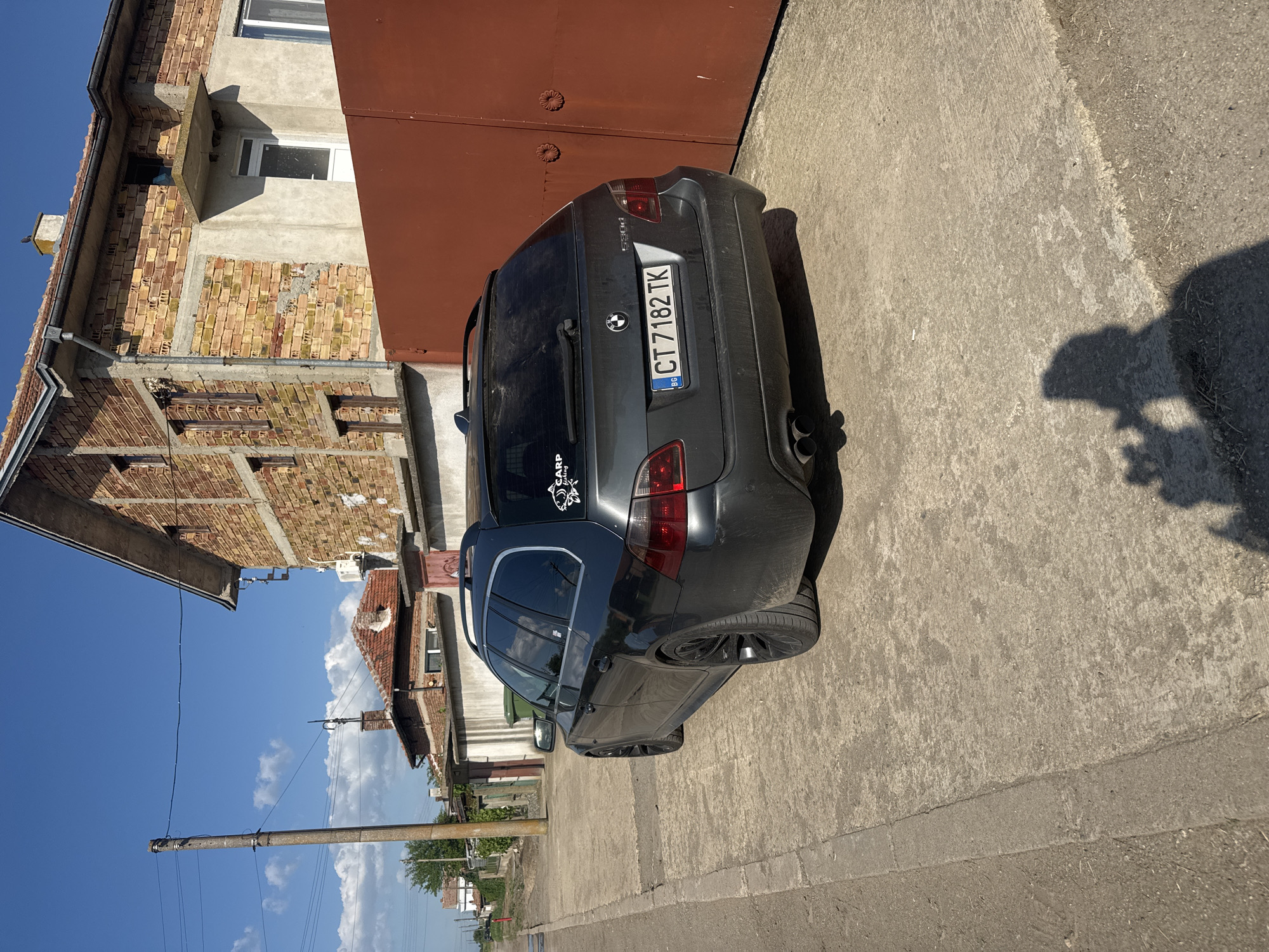 The image depicts a grey BMW 530d station wagon parked on a concrete surface next to a building in Sarnevo, Bulgaria. The car is positioned on the right side of the frame, with its rear end facing the viewer. The license plate number is CT 7182 TK. A sticker is visible on the rear window. A building with a combination of brick and concrete construction stands to the left of the car. Adjacent to the car is a large reddish-brown garage door. The concrete surface appears to be a sidewalk or a paved area. There is a shadow of a person present, and in the background, there are utility poles, wires, trees, and other buildings. The sky is blue with some clouds. The location is likely a residential street in Sarnevo, Bulgaria.