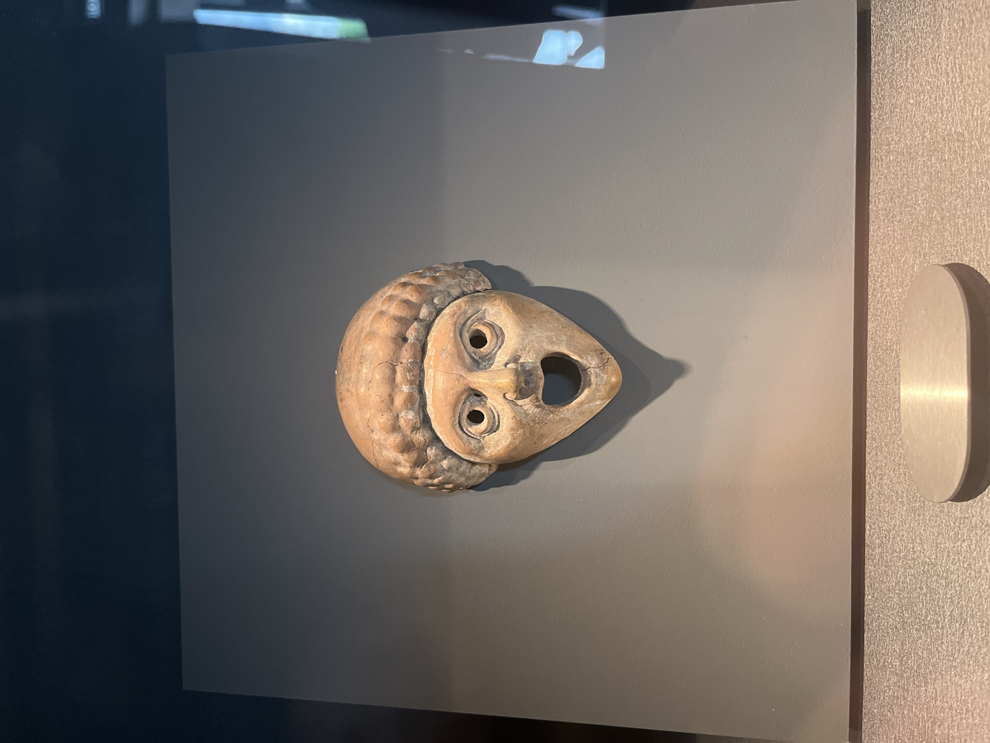A terracotta owl mask is displayed on a gray background. The mask has a rounded head with textured, feather-like details around the edge. Two circular eye holes are visible above a triangular nose opening. A small, dark indentation suggests a beak. The mask is slightly tilted and casts a shadow on the gray surface. The scene is likely in a museum or exhibit, as indicated by the presentation. A white, rounded fixture is visible on the right edge of the image. The location details (city and country) are unknown from the image.