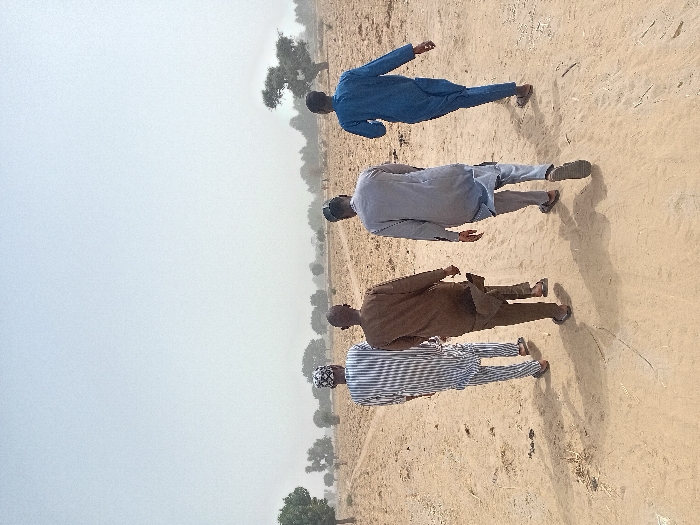 Four adult males are observed walking from a rear perspective along a dusty dirt path in Ayama, Nigeria. The environment is an open, arid landscape under a hazy sky.
The individuals are dressed in loose-fitting traditional attire and open-toed sandals:
- The leftmost man wears a white and blue striped long-sleeved tunic and matching trousers, topped with a patterned cap.
- The second man from the left wears a solid brown long-sleeved tunic and trousers.
- The third man wears a light grey long-sleeved tunic and trousers.
- The rightmost man wears a solid blue long-sleeved tunic and matching trousers.
All four individuals are walking in the same general direction, casting shadows forward and to their left on the light brown earth. The path appears unpaved with visible tracks. The surrounding terrain is characterized by dry, light brown soil with sparse, low-lying vegetation and scattered deciduous trees in the mid-ground and background. The sky is a pale, uniform light grey, indicative of hazy or overcast conditions.