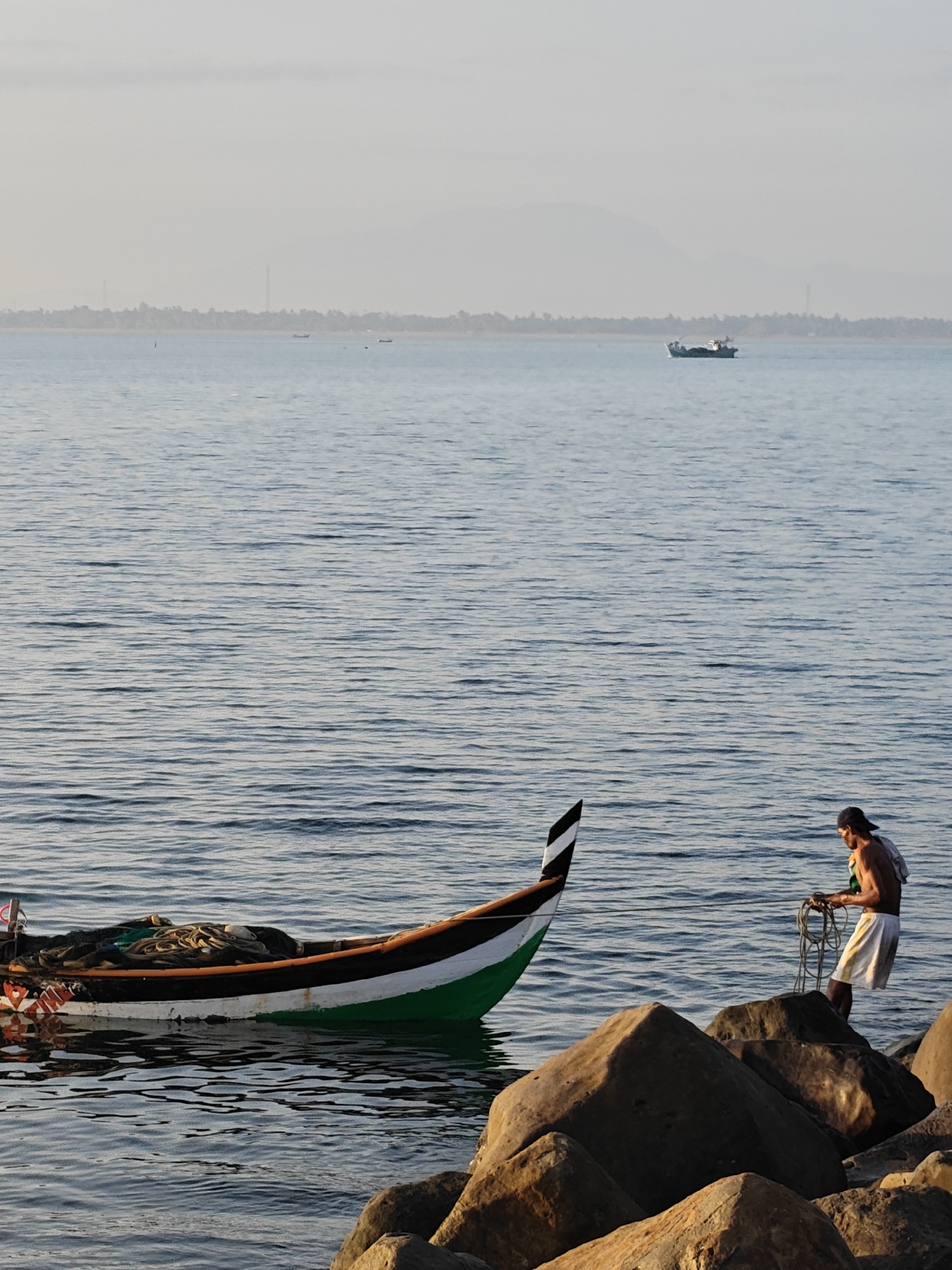 In the city of Lhokseumawe, Indonesia, at the edge of the sea, a man stands on rocks near the water. He is wearing a dark baseball cap, white shorts, and appears to be shirtless. He holds a coil of rope in his hands, seemingly preparing to untangle or manage it. To his left, a small, narrow boat rests on the water, its hull painted green and white with a black stripe along the top. The boat is laden with fishing nets. The sea is calm with gentle ripples. In the background, the hazy outline of distant land with trees and structures is visible, along with several other boats on the horizon. The sky is pale and overcast, suggesting either early morning or late afternoon light. The foreground is dominated by large, textured rocks, casting shadows.