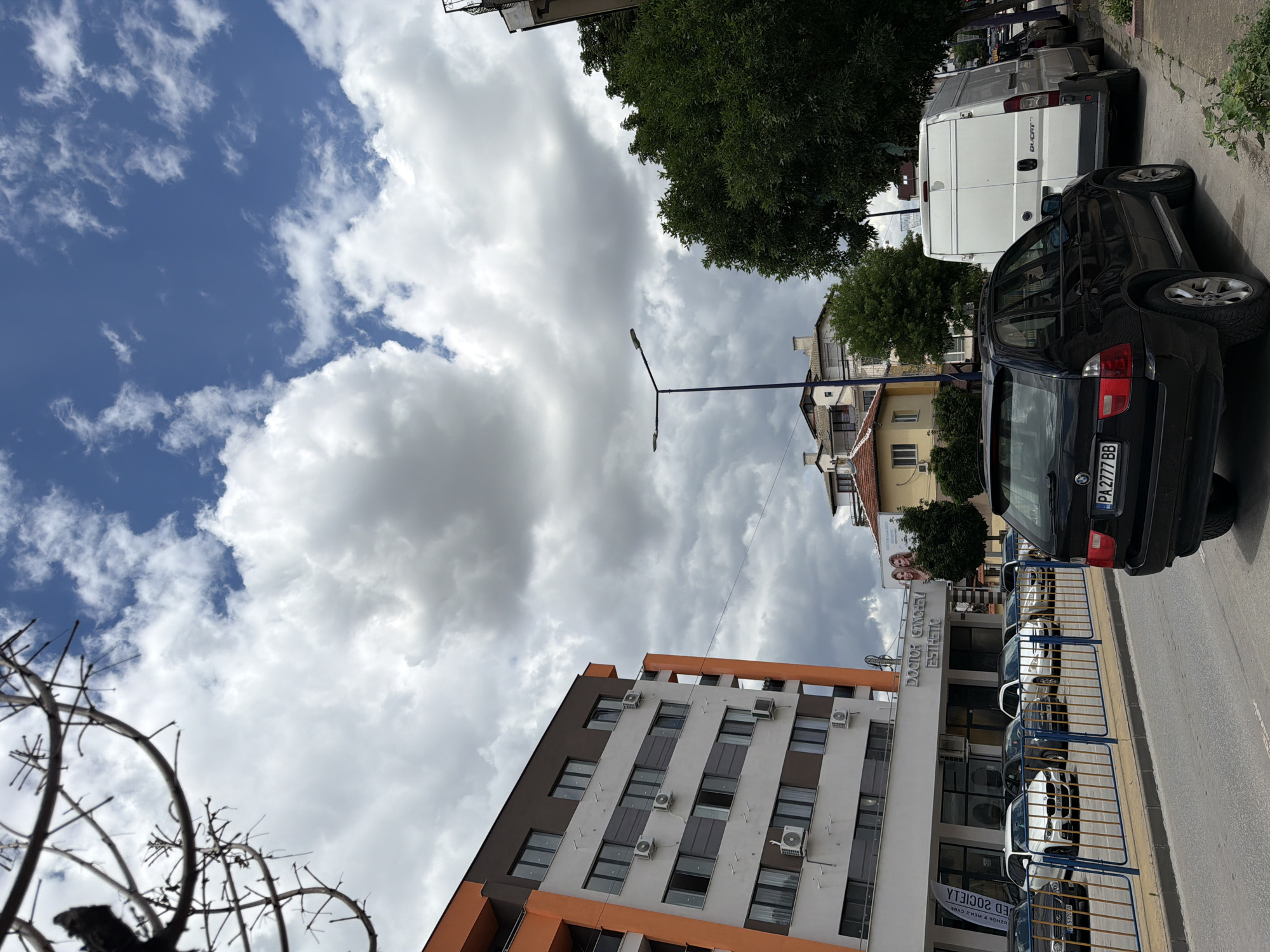 The image depicts a street scene in Pazardzhik, Bulgaria, with a modern apartment building and several vehicles. The apartment building, a multi-story structure with orange and brown accents, features numerous windows and air conditioning units. In front of the building, a row of cars is parked along the sidewalk, including a black BMW X5 SUV and a white van. The street is lined with trees, and the sky is filled with dramatic, fluffy white clouds against a blue backdrop. The presence of "DOCTOR GANCHEV ESTHETIC" signage on the building suggests a commercial establishment on the ground floor. A street lamp is visible in the mid-ground.
