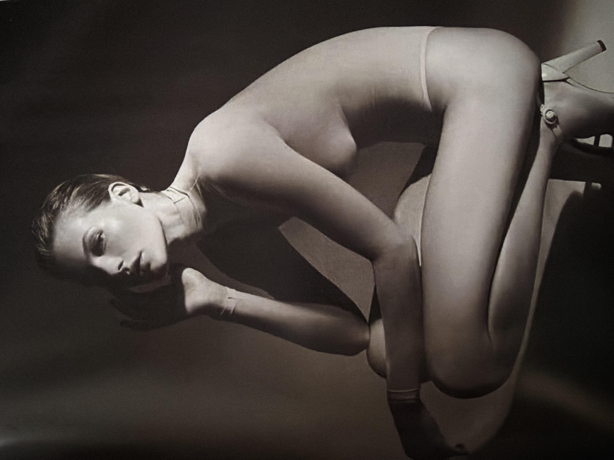A black and white artistic photograph shows a nude woman in a contorted pose, with her body arched and her head tilted towards the viewer. She is wearing a high-heeled shoe on one foot. The background is dark and unclear. The photograph is taken indoors. No geographical location is identifiable.