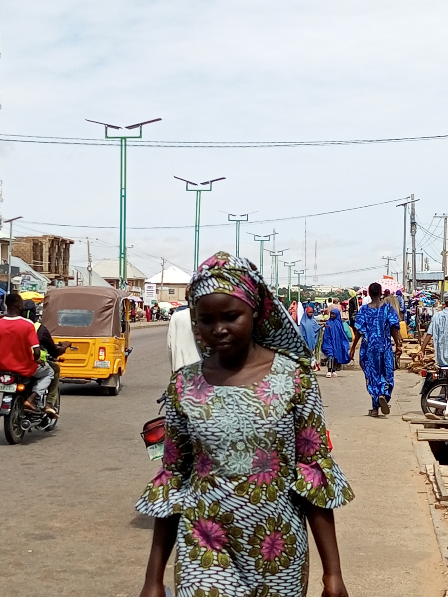 The image depicts a street scene in Tempure, Nigeria, on a cloudy day. In the foreground, a young woman walks towards the camera, wearing a colorful patterned dress and headscarf. The dress features floral motifs in pink and green against a white and black striped background. Behind her, the street is bustling with activity. A yellow auto-rickshaw (Keke Napep) is visible to her left, with a person in a red shirt on a motorcycle beside it. Further down the street, several people are walking, including a woman in a vibrant blue dress. The environment includes a paved road, sidewalks lined with market stalls, and streetlights with solar panels. The sky is overcast with some visible power lines. The scene captures the everyday life and commerce of a Nigerian city.