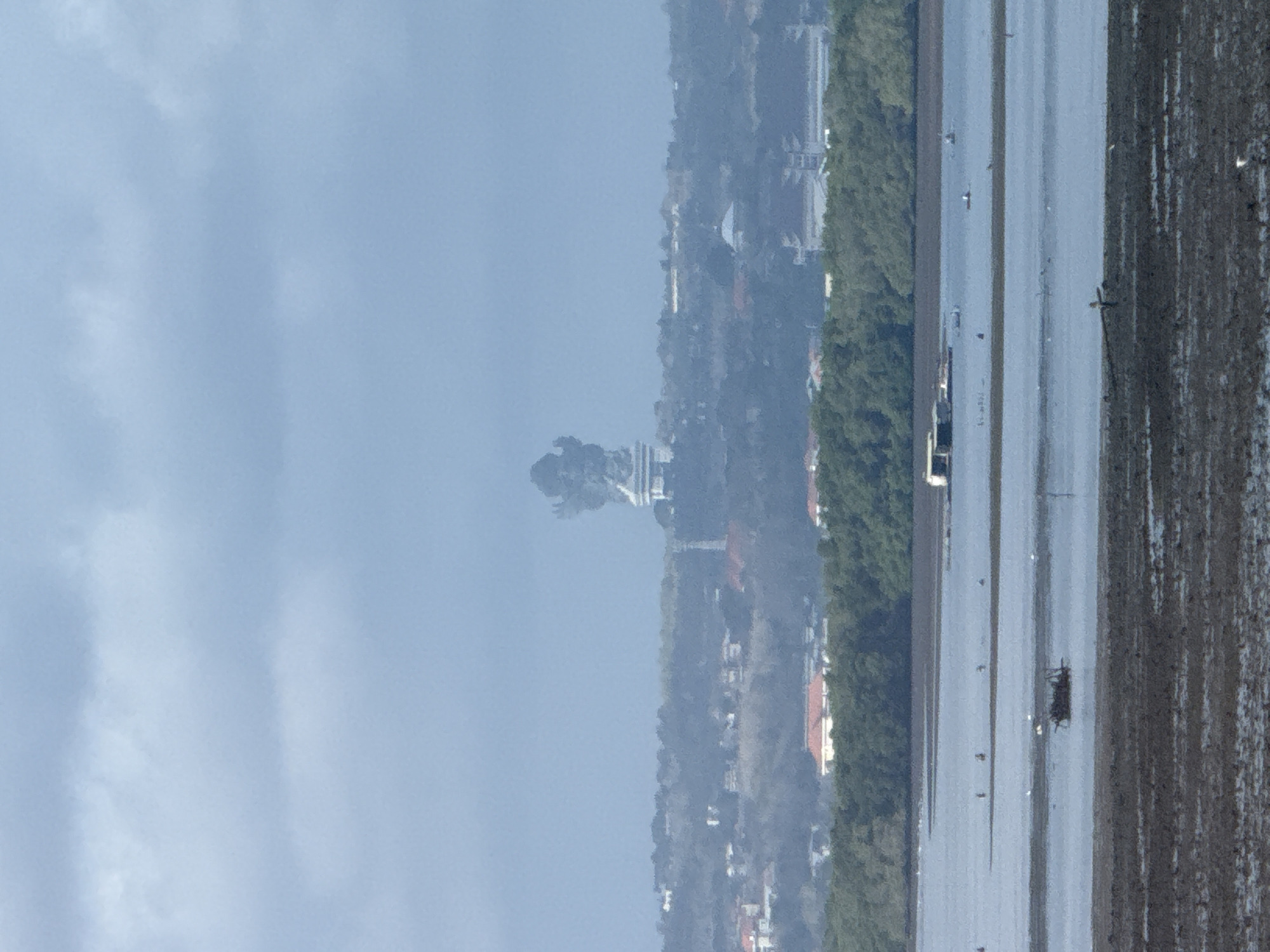 The image depicts a hazy, overcast day with a wide expanse of water in the foreground, likely a bay or inlet, with a muddy shoreline. In the middle ground, a lush green line of trees separates the water from a cityscape. The city features buildings with red-tiled roofs and other structures scattered amidst the greenery. Dominating the background skyline, partially obscured by atmospheric haze, is a large, dark, monolithic statue or monument, possibly of religious or cultural significance. A few small boats or floating structures are visible on the water. The overall impression is one of a distant, somewhat obscured view of a coastal settlement with a prominent landmark. The location is identified as Garuda Wisnu Kencana Cultural Park, Bali, Indonesia, famous for its colossal statue.