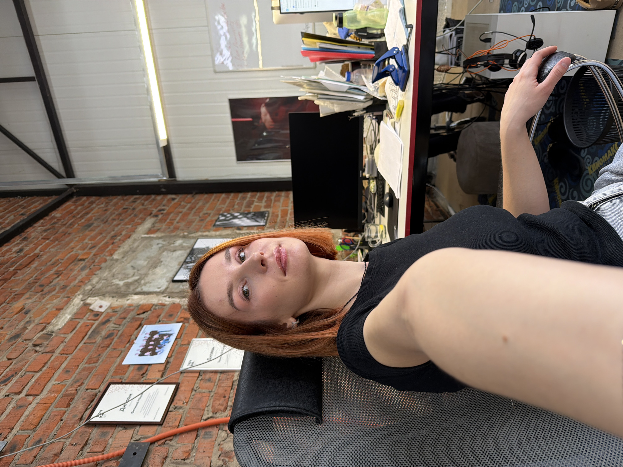 The image is a selfie taken in what appears to be an office or studio setting in Chaikovsky, Russia, based on visible wall art and possibly a diploma. The person is seated in an office chair, with a black sleeveless top and reddish-brown hair. The background includes a brick wall with framed pictures or documents, a cluttered desk with a monitor and office supplies, and a partially visible ceiling with metal beams.