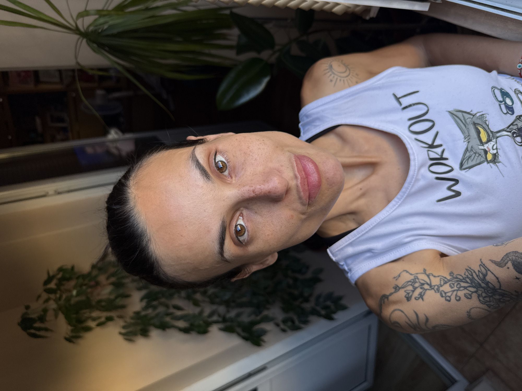 A woman with brown eyes and dark hair pulled back poses for a selfie in what appears to be an apartment balcony. She is wearing a white tank top with the word "WORKOUT" printed on it and a cartoon cat graphic. She has several tattoos on her arms, including a floral design and a sun and moon symbol. Plants are visible in the background, suggesting an outdoor or semi-outdoor setting. The lighting suggests it is daytime. The location is identified as Pleven, Bulgaria.