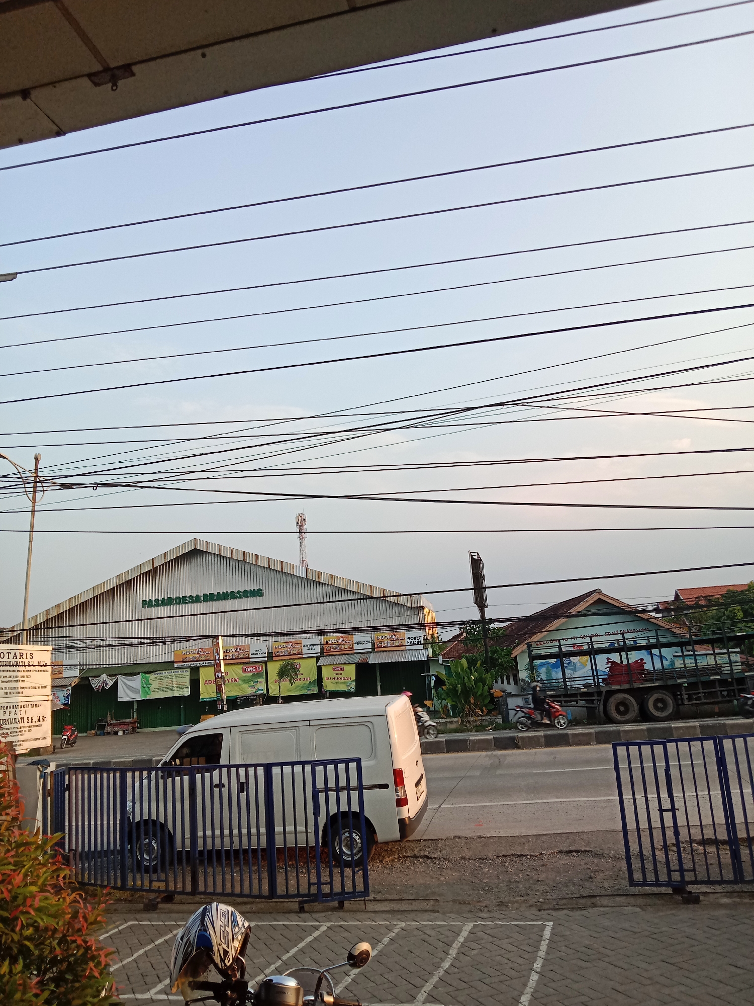 The image shows a street scene in Brangsong, Indonesia, with a large market building in the background. The market, identified by the sign "PASAR DESA BRANGSONG," has a distinctive peaked roof. In front of the market, there are several banners and advertisements, suggesting a vibrant commercial area. A white van is parked on the side of the road, and a blue metal fence runs along the foreground. A motorcycle helmet rests on the handlebars of a visible motorcycle in the lower part of the image. A truck with a loaded bed is also visible further down the road. The sky is clear with a few wispy clouds, and power lines crisscross the scene. The overall impression is of a typical day in a bustling Indonesian town.