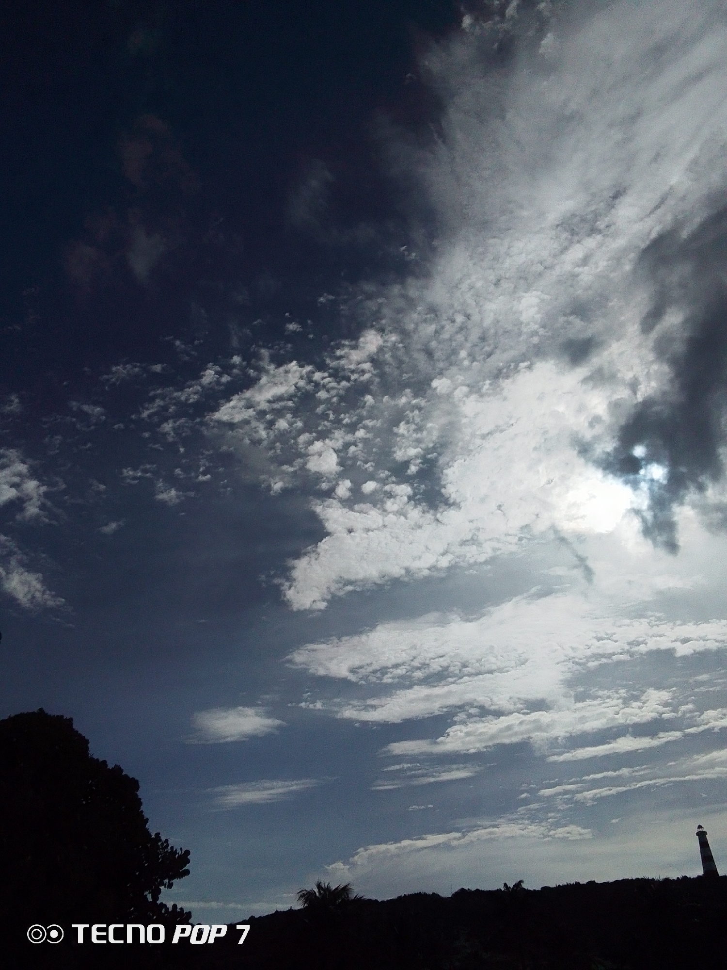 The image depicts a cloudy sky in Pedro González, Venezuela. The sky is dominated by dark blue and white clouds, with the sun partially visible through a break in the clouds on the right side. The horizon is silhouetted, with the outlines of trees, vegetation, and a lighthouse discernible. The bottom left corner shows a dark tree. The words "TECNO POP 7" are displayed in the bottom left corner of the image.