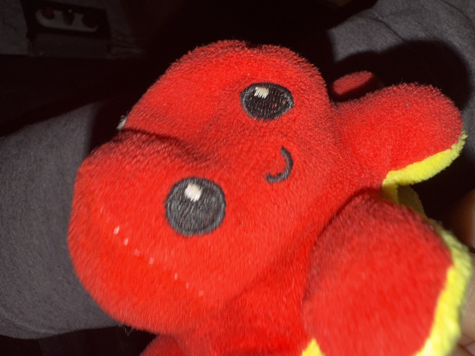 The image depicts a close-up, indoor scene in Bakurtsikhe, Georgia, focusing on a red plush reversible octopus toy. The toy, positioned prominently in the foreground and center, features black embroidered eyes with white pupils and a black embroidered smile. Its underside or reverse side, visible on the tentacles, is yellow. The red plush surface of the toy shows several small, light-colored particles, consistent with lint or dust. The toy is actively held by an unseen individual; a portion of a human hand is visible at the lower left of the frame. The background is dark and heavily out of focus, with a section of dark grey fabric, possibly clothing worn by the individual, visible behind the toy. A faint, indistinct light-colored shape is discernible in the upper right quadrant of the dark background. The scene is illuminated by direct, localized lighting, emphasizing the toy while rendering the background largely obscure.