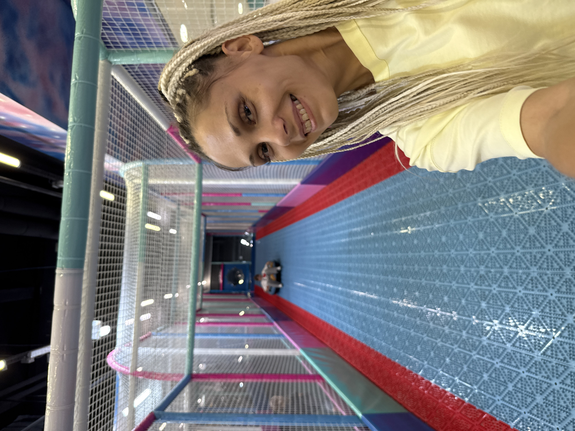 A woman with blonde braids is taking a selfie in a brightly colored indoor play area. The play area has blue, red, and pink sections, with nets and tubes forming tunnels and slides. In the background, a child is sliding down a tube in a sled. The woman is smiling and wearing a yellow t-shirt. The location is identified as Rusanovo, Russia.
