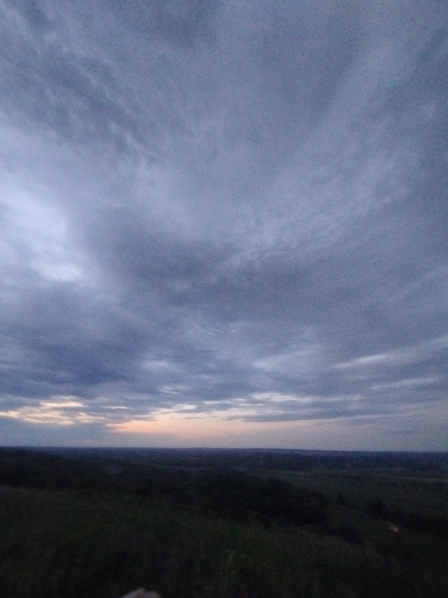 The image shows a wide shot of a dusky landscape. The sky is filled with a mosaic of grey clouds, with hints of pale pink and orange near the horizon, suggesting either sunrise or sunset. Below the sky, a dark, rolling landscape stretches out, appearing to be covered in vegetation, possibly fields or forests. The foreground is dark and appears to be a grassy hill. There are no visible people or objects in the scene. The location is specified as Unknown, Unknown.