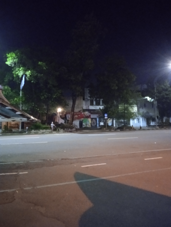 The image shows a street in Surakarta, Indonesia, at night. The street is empty, except for a single person walking in the distance. The road is asphalt and has a slight incline. There are white painted lines running across the width of the road. On either side of the road are trees, bushes, and a building with faded graffiti on its exterior walls. The image was captured from the perspective of a person standing on the sidewalk. The building is located at the coordinates -7.569 latitude, 110.82 longitude. The light is dim and there are street lights in the distance. The image is grainy, which could be a result of low light conditions. There are no cars or other vehicles on the street. The trees obscure the view of the sky, but it appears to be clear. The building on the left side of the road is painted in shades of white and gray, and has a simple, flat roof. There are no visible doors or windows on the building. The building on the right side of the road appears to be made of concrete or brick, and has a more complex facade. The building is decorated with graffiti and faded advertisements, but the details are obscured by the darkness. There are trees on both sides of the street, but they are not in focus. They are all densely packed, suggesting that it is a heavily populated area. The trees are all large and mature. The sidewalk appears to be made of concrete and is wide enough for pedestrians to walk on. There is a long, narrow, gray building behind the trees. The building has a flat roof and seems to be made of concrete. The building has a window on the front and a faded sign that is not visible. The image is taken from a slightly elevated position, which gives the viewer a good view of the street and the buildings. There are no other people visible in the scene. The atmosphere of the image is one of stillness and peacefulness. The lack of activity on the street adds to the sense of calm.