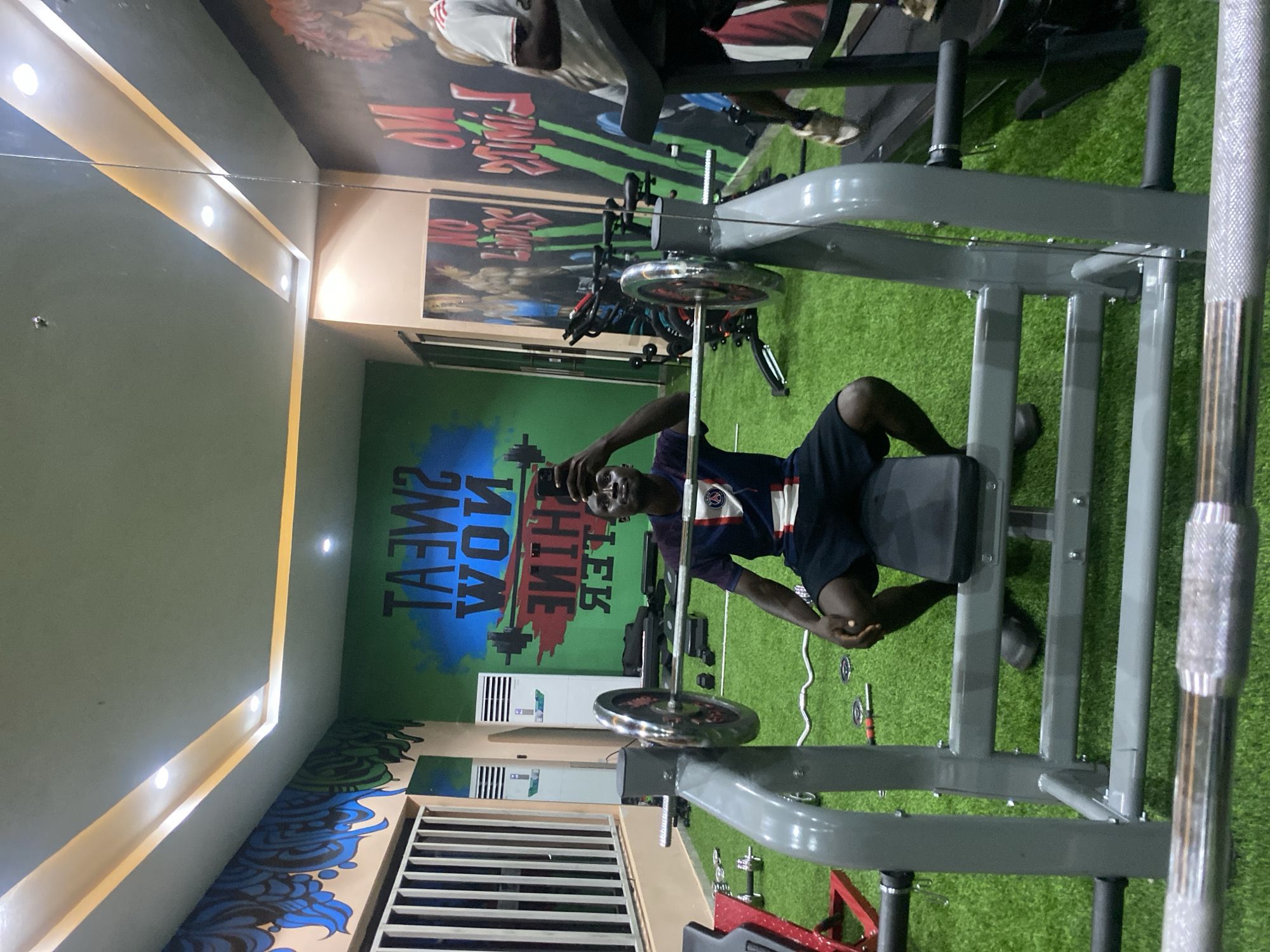 A young Black man is taking a selfie in a gym in Barden Yana, Nigeria. He is seated on a weight bench with a barbell in front of him. He is wearing a blue and white striped t-shirt and black shorts. The gym has a green artificial turf floor, and the walls are decorated with motivational murals. There are various weightlifting machines and equipment visible in the background. The lighting in the gym is provided by ceiling lights and accent lighting strips.