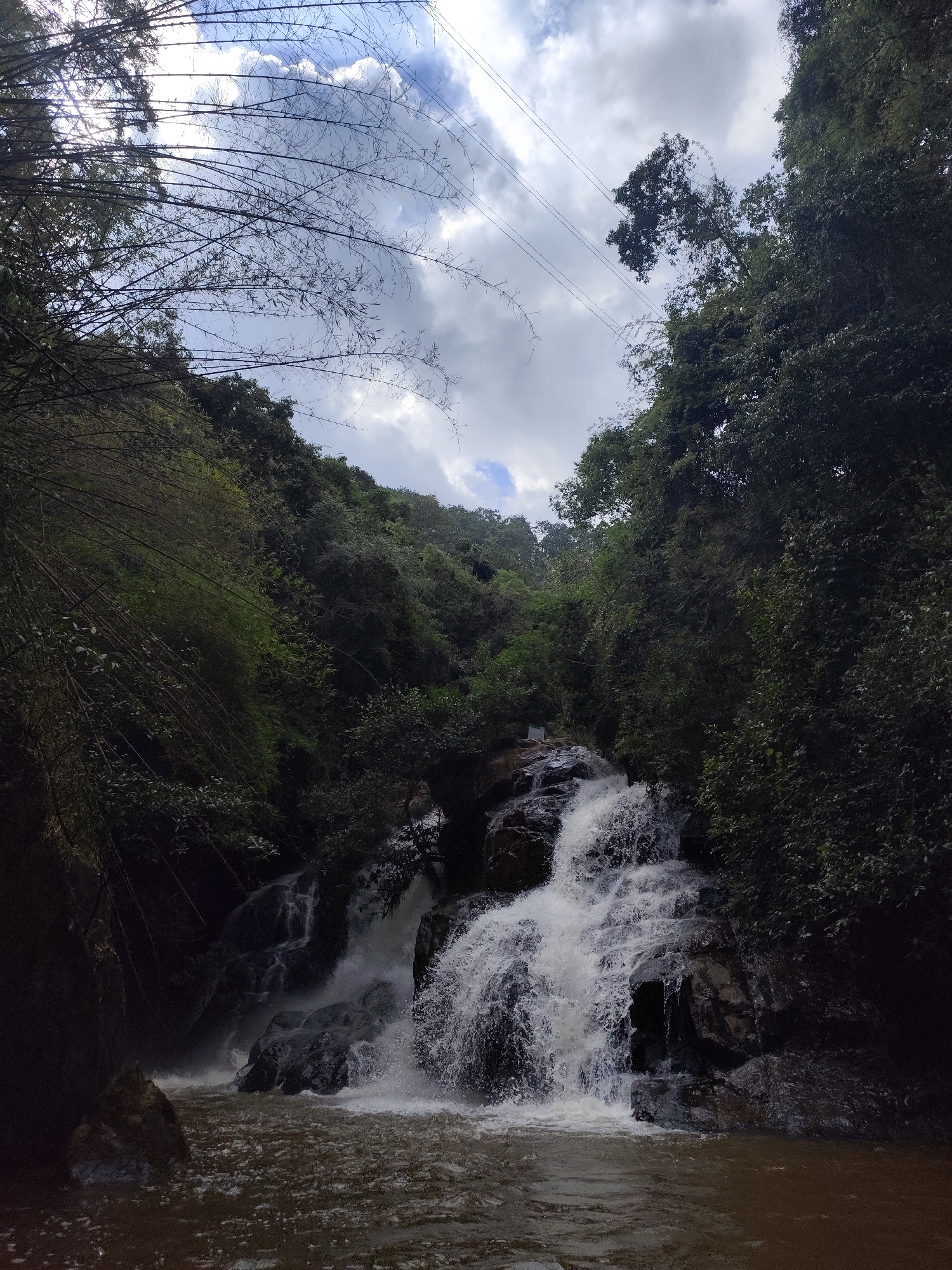 The image shows a cascading waterfall in a lush, green environment under a cloudy sky. The waterfall is the focal point, with water rushing over dark rocks into a pool below. Vegetation, including bamboo and various trees, frames the scene. Power lines are visible in the upper portion of the image. The location is likely near Da Lat, Vietnam.