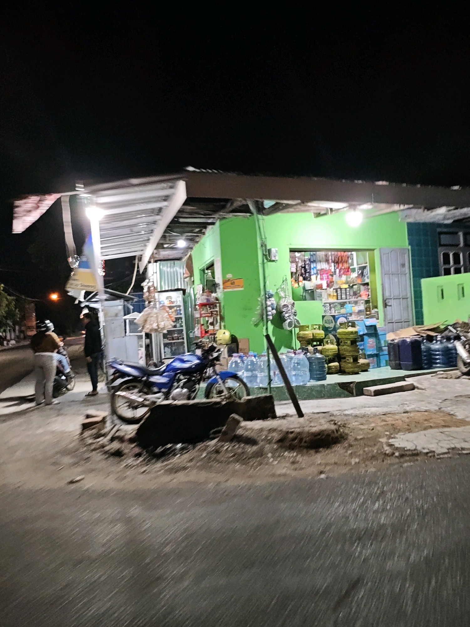 The image depicts a street scene at night in Pagu, Indonesia, likely in front of a small store. A bright green building, possibly a shop, is illuminated by fluorescent lights. In front of the store, there are stacks of large water bottles and propane gas cylinders. A blue motorcycle is parked on the side, and another person on a motorcycle is visible further down the street. The ground is paved with asphalt and concrete, with some dirt and debris scattered around. The overall atmosphere is dimly lit and appears to be a typical evening in a residential or commercial area.