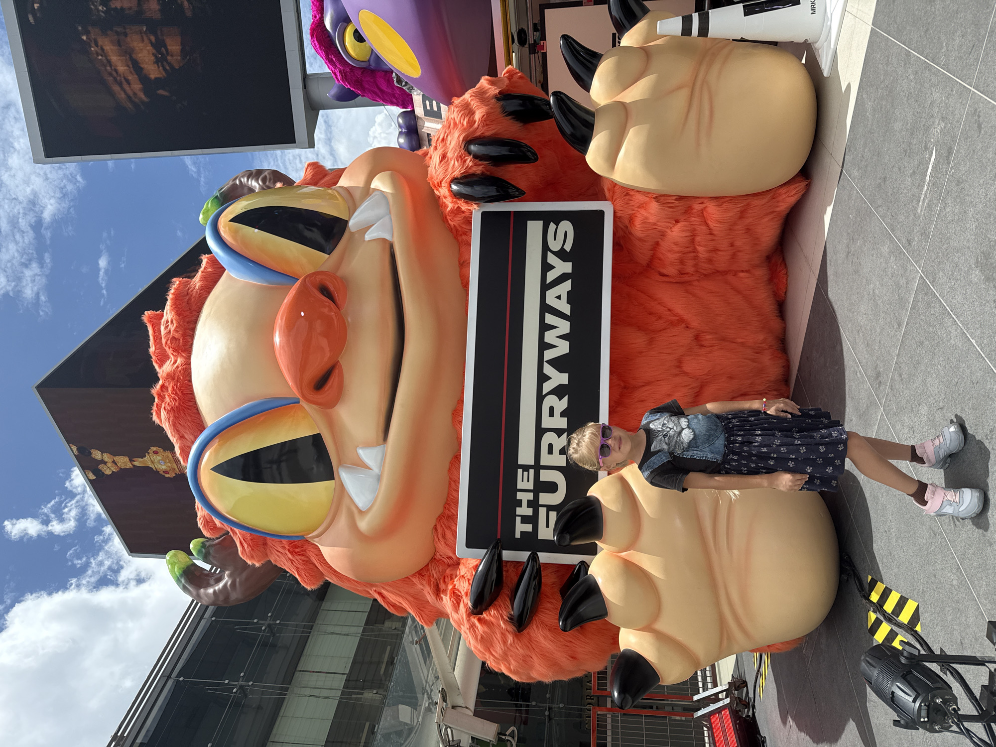 A young girl with blonde hair, wearing sunglasses, a t-shirt with a cat graphic, and a dark blue patterned dress over overalls, stands to the right of a large, fluffy orange monster prop. The monster has large, yellow eyes with blue accents and sharp white teeth. It is holding a black sign with white text that reads "THE FURRWAYS." The scene appears to be outdoors in a city setting, with buildings and a large screen visible in the background. The ground is paved with grey tiles. The girl is looking at the camera. The overall impression is of a promotional event or attraction, likely in Bangkok, Thailand, given the context provided.