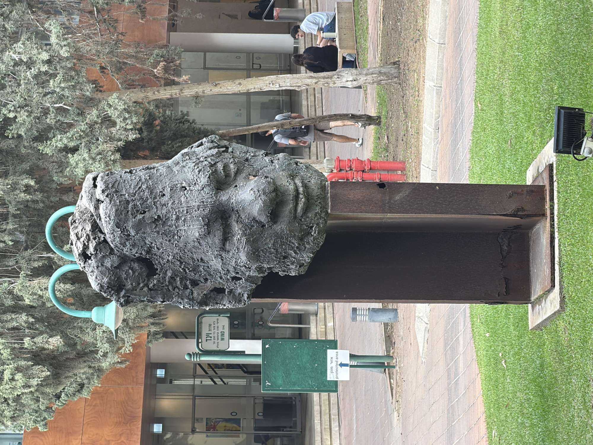 The image shows a large, rough sculpture of a human head mounted on a rusty steel structure, outdoors. The statue is located in Herzliya, Israel as indicated by the street sign that reads "Wind Blvd." in Hebrew and English, a street named after Prof. Gidi Arnsan Wind, one of the founders of the Interdisciplinary Center Herzliya. Two street lamps with curved arms are positioned near the sculpture. A sign that says "Protected Area" in Hebrew and English is also visible. In the background, people are walking or sitting on a bench. The area is partially paved with bricks and partially covered with green grass. Trees surround the statue.