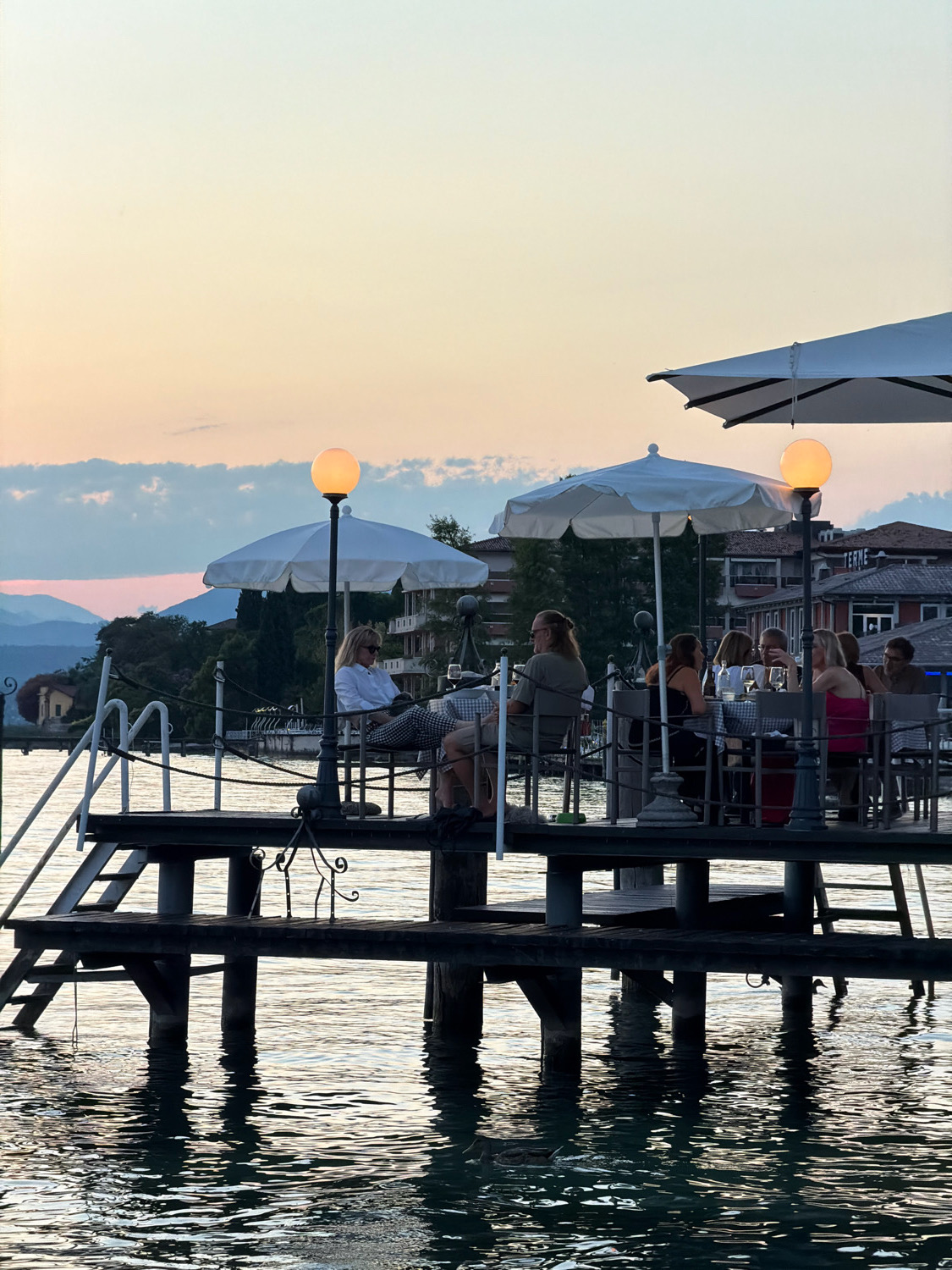 This image depicts a waterside dining scene at sunset, likely in Sirmione, Italy, given the distinctive architecture visible in the background. A wooden pier extends into the calm water, supporting a restaurant or cafe area. Several groups of people are seated at tables, enjoying the evening. White umbrellas provide shade for the diners, and illuminated lanterns cast a warm glow. The sky is painted with soft hues of orange, pink, and yellow, characteristic of a sunset over a body of water. The water itself reflects the sky and the structures on the pier, creating a serene and inviting atmosphere. Buildings with red-tiled roofs and a few trees are visible in the distance, adding to the picturesque setting. The presence of a duck swimming in the foreground suggests a natural and relaxed environment.