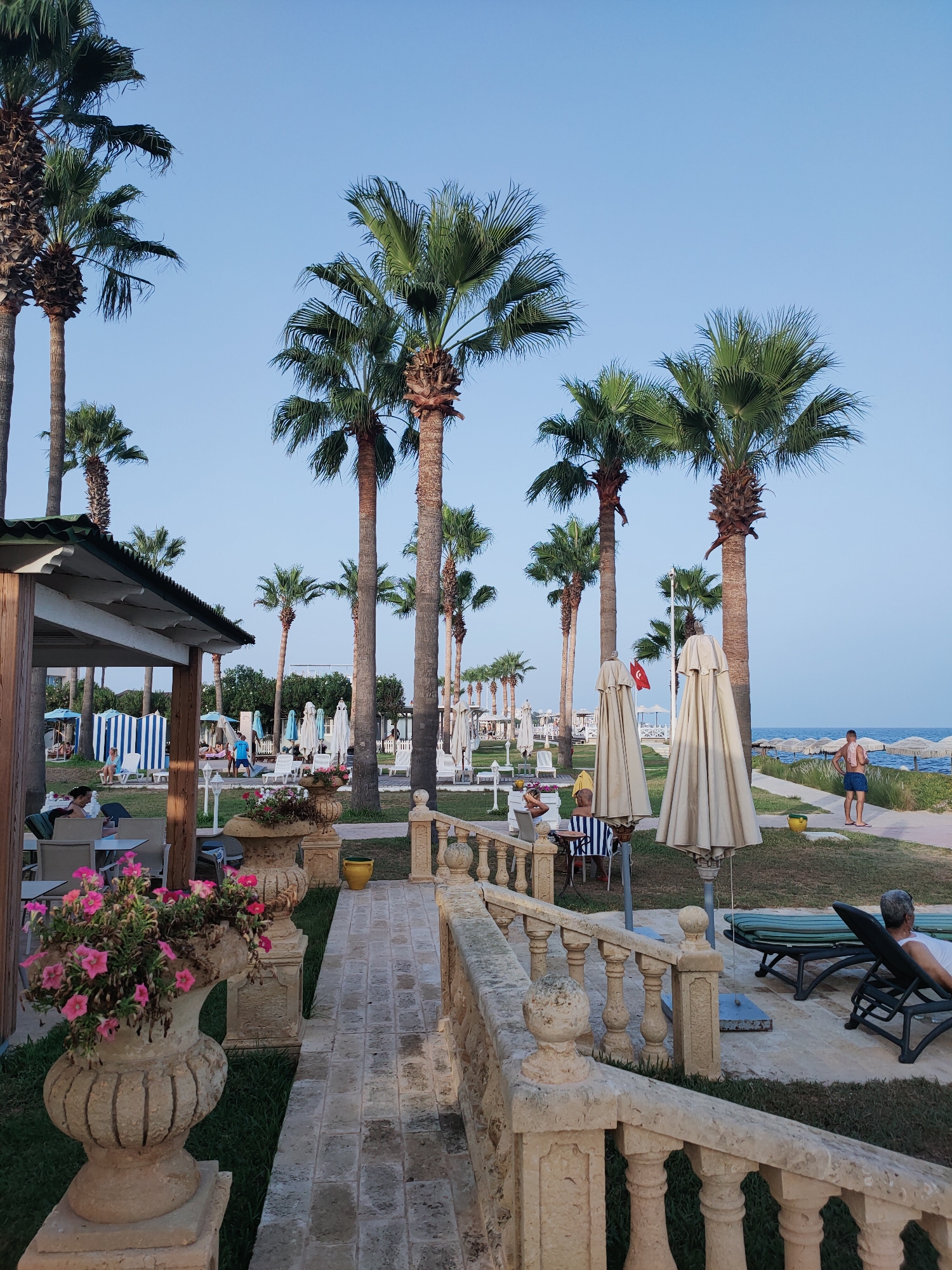 The image depicts a resort area with palm trees and beach chairs. The scene is set in Nabeul, Tunisia, indicated by a Turkish flag visible in the background. A man in blue shorts stands near the sea, facing away from the camera. In the foreground, a stone railing overlooks a walkway and a garden with flowers in decorative pots. A covered seating area with tables and chairs is visible to the left, with a few people seated. Several closed umbrellas and lounge chairs are scattered across the grass and paved areas. The sky is clear and blue.