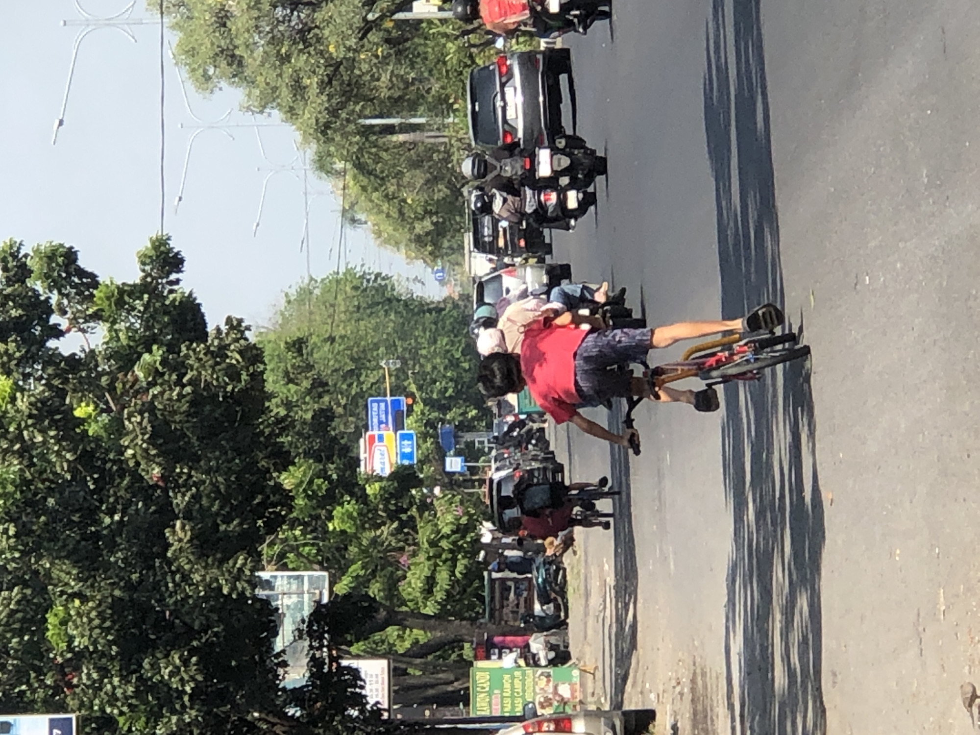 The image depicts a street scene in Larangan, Indonesia, on a sunny day. In the foreground, a young boy wearing a red t-shirt and patterned shorts cycles a red and black bicycle down the asphalt road. His legs are visible, one pedaling, and he is wearing dark sandals. In the midground, two other children on a bicycle, one in a red shirt and the other in a darker shirt, are riding ahead of him. Several vehicles, including motorcycles, cars, and a van, are present on the road, some of which are carrying passengers. On the left side of the street, there are several shops or stalls with signs, and lush green trees line the road. Streetlights and utility poles are visible in the background, indicating an urban or suburban setting. The overall atmosphere is that of a typical street scene with active traffic and pedestrian activity.