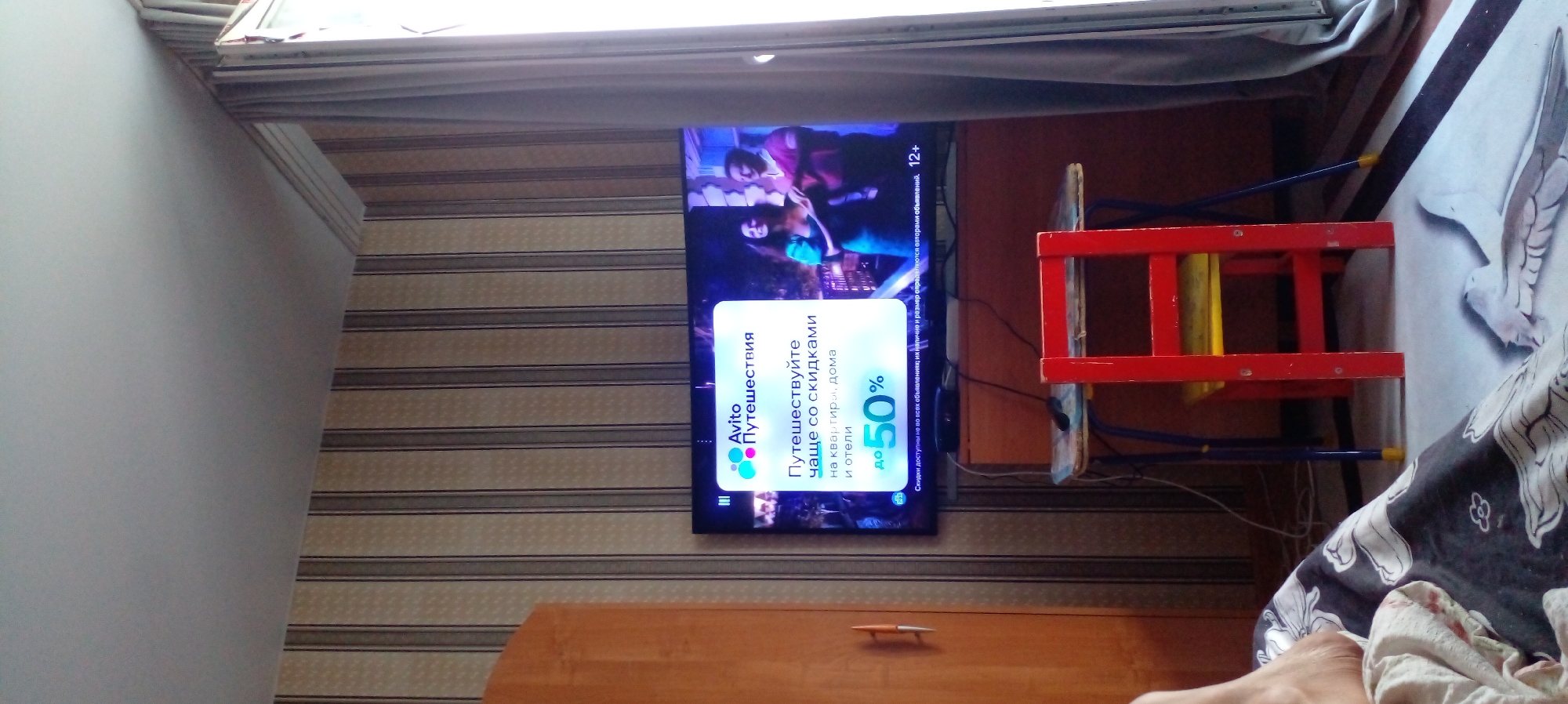 The image shows an interior scene with a television screen displaying an advertisement for Avito "Travel" services, featuring a discount of up to 50%. The TV is situated on a desk with a red chair in front of it. To the left of the TV, a wooden wardrobe is visible, and to the right, a grey curtain hangs. The walls are covered with striped wallpaper. The scene appears to be a living room or a bedroom. There are no individuals present in the immediate foreground or background of the shot. The image's context does not provide any specific information to determine its location within the city of Glubokoe, Kazakhstan.