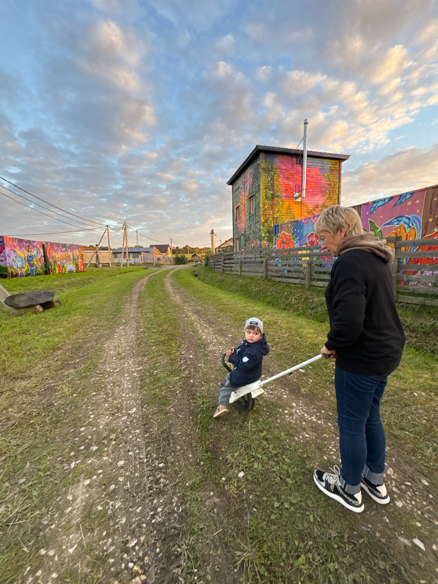 In Kempelevo, Russia, a woman with blonde hair is pushing a young child sitting on a toy vehicle. The child is wearing a gray cap and a dark blue jacket. The woman is wearing a black hoodie, blue jeans, and black and white sneakers. They are on a dirt path in a grassy area, with power lines and a colorful building in the background. The building is painted with bright murals of trees and abstract designs, and a metal chimney extends from its roof. A wooden fence runs along the side of the path, separating the road from more colorful murals depicting figures and flowers. The sky is filled with clouds, with hues of orange and pink indicating either sunrise or sunset.