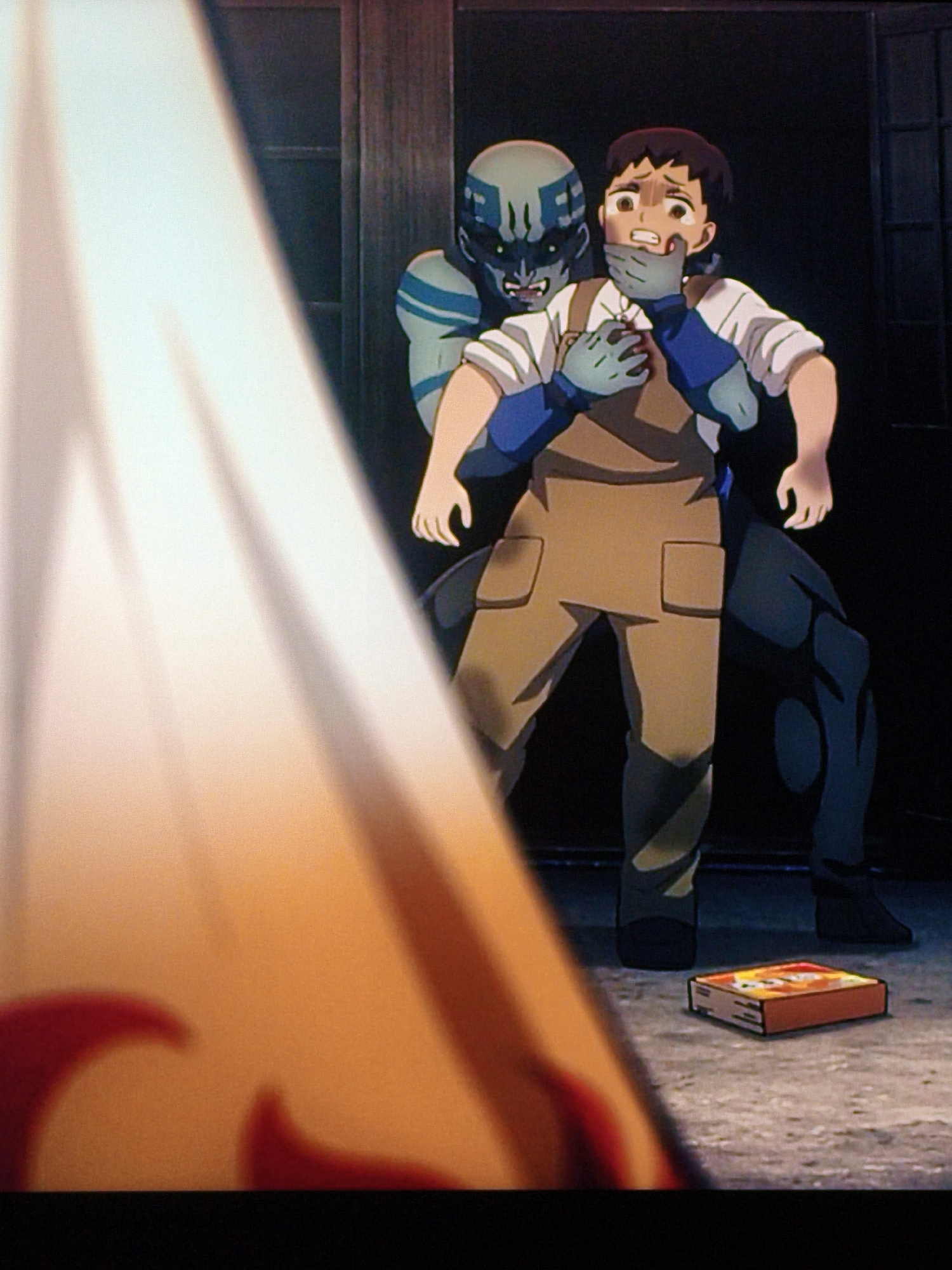 An animated scene depicts a young person with short dark hair and wide eyes, wearing a white shirt and a brown apron, being held from behind by a blue-skinned creature with dark markings on its face and chest. The creature's mouth is open, revealing sharp teeth, and its hand is clamped over the young person's mouth. The young person's arms are held down by the creature. In the foreground, a large, out-of-focus object with red markings is visible. On the ground in the background, a small, rectangular object resembling a box of matches or crackers is present. The environment appears to be indoors, with wooden structures and dark walls. The lighting suggests a dimly lit interior with a strong light source casting shadows. The scene does not contain any specific geographical markers that would identify it as being located in Vladivostok, Russia.